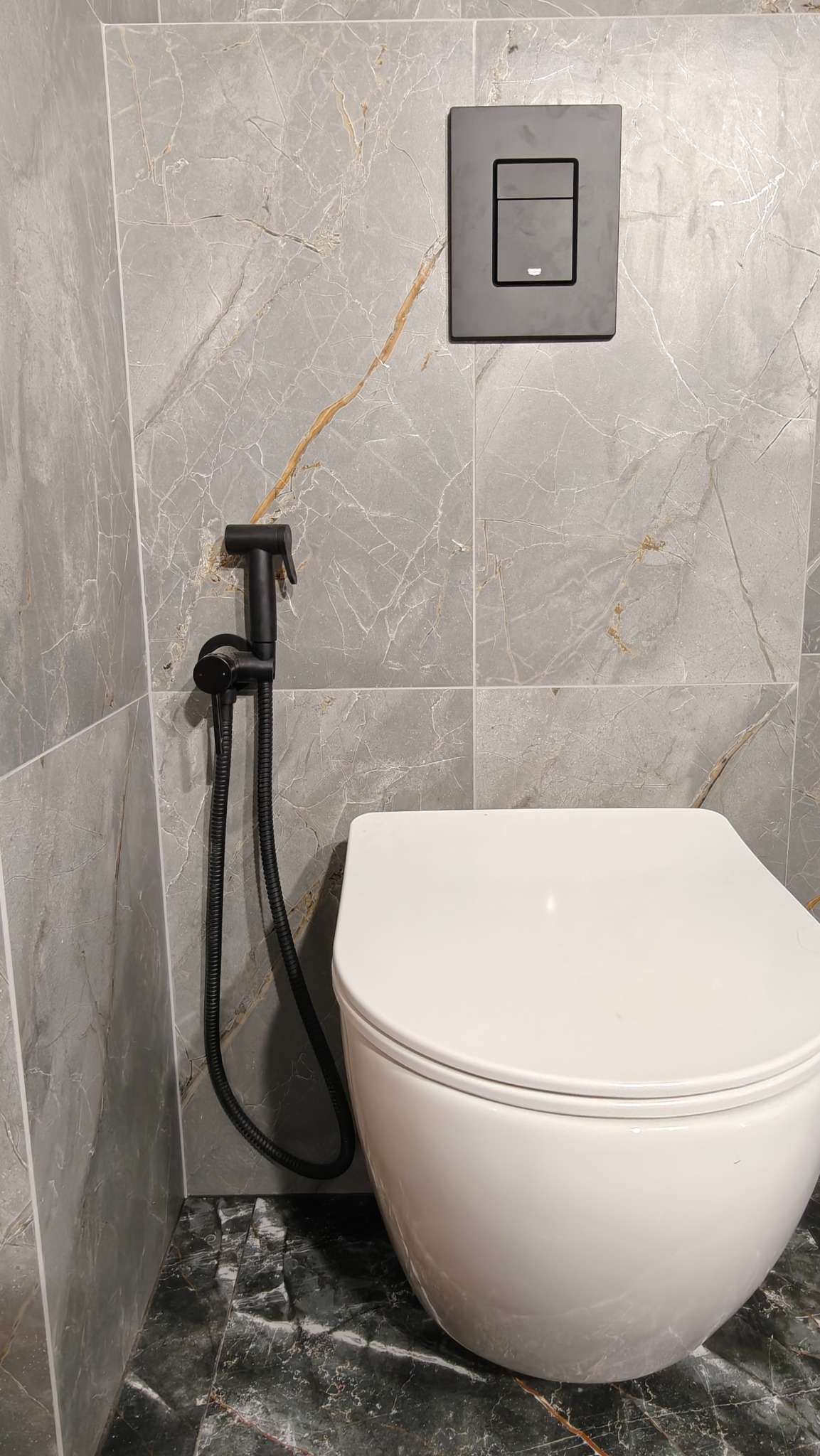 Nowoczesna toaleta podwieszana z bidetką w kolorze czarnym, na tle szarych płytek imitujących marmur z złotymi żyłami. Widoczny czarny przycisk spłuczki.