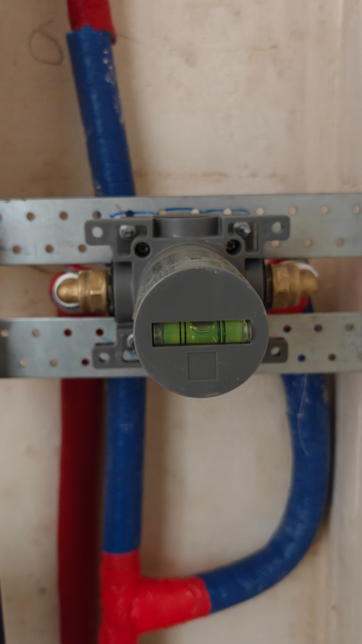 Szary rozdzielacz z poziomnicą, zamontowany na metalowej listwie, z podłączonymi rurami w kolorze niebieskim i czerwonym, na tle jasnej ściany.