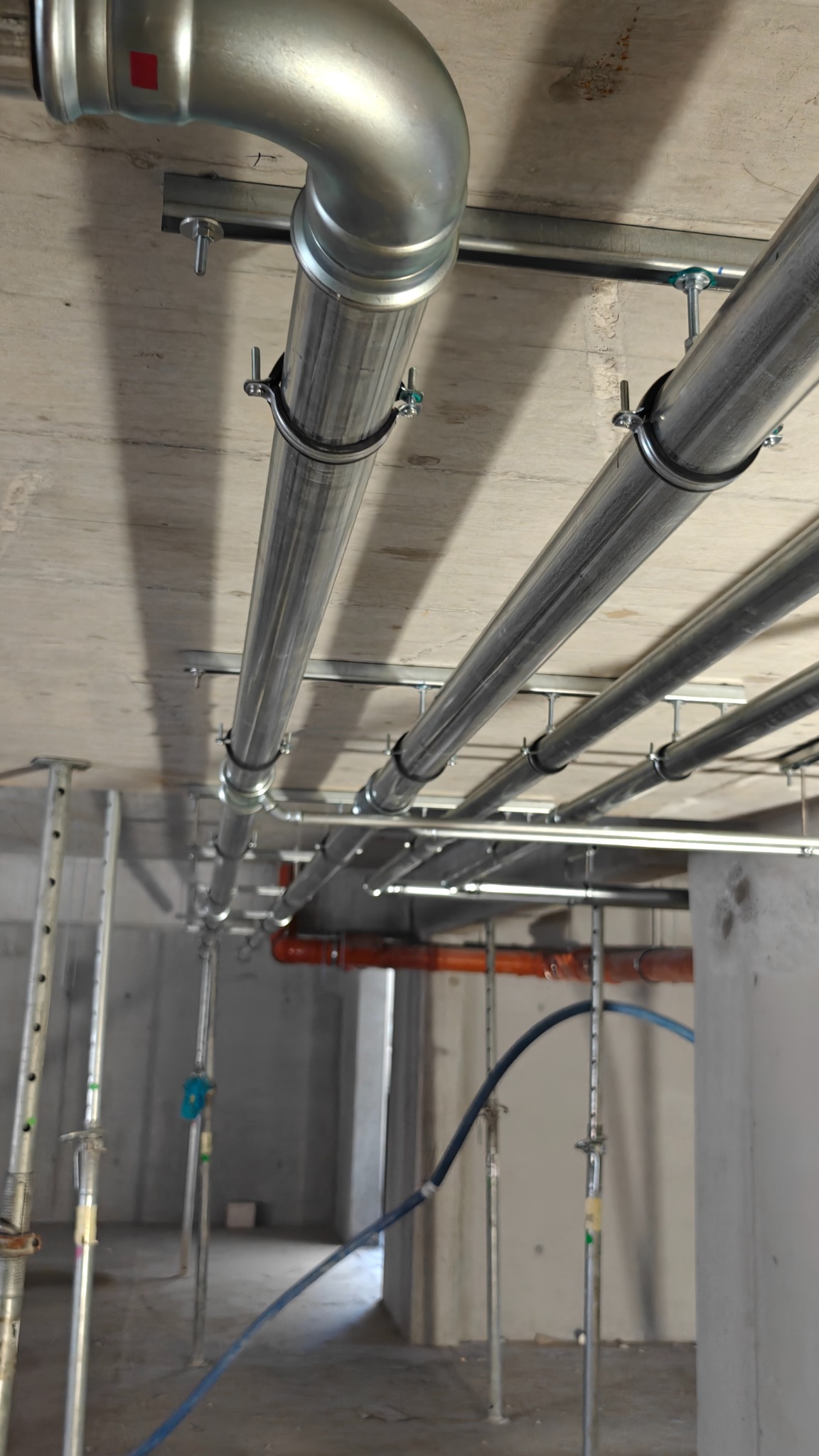 Metalowe rury instalacyjne zamocowane do sufitu w surowym wnętrzu. Widoczne wsporniki i obejmy. Część instalacji w kolorze pomarańczowym.
