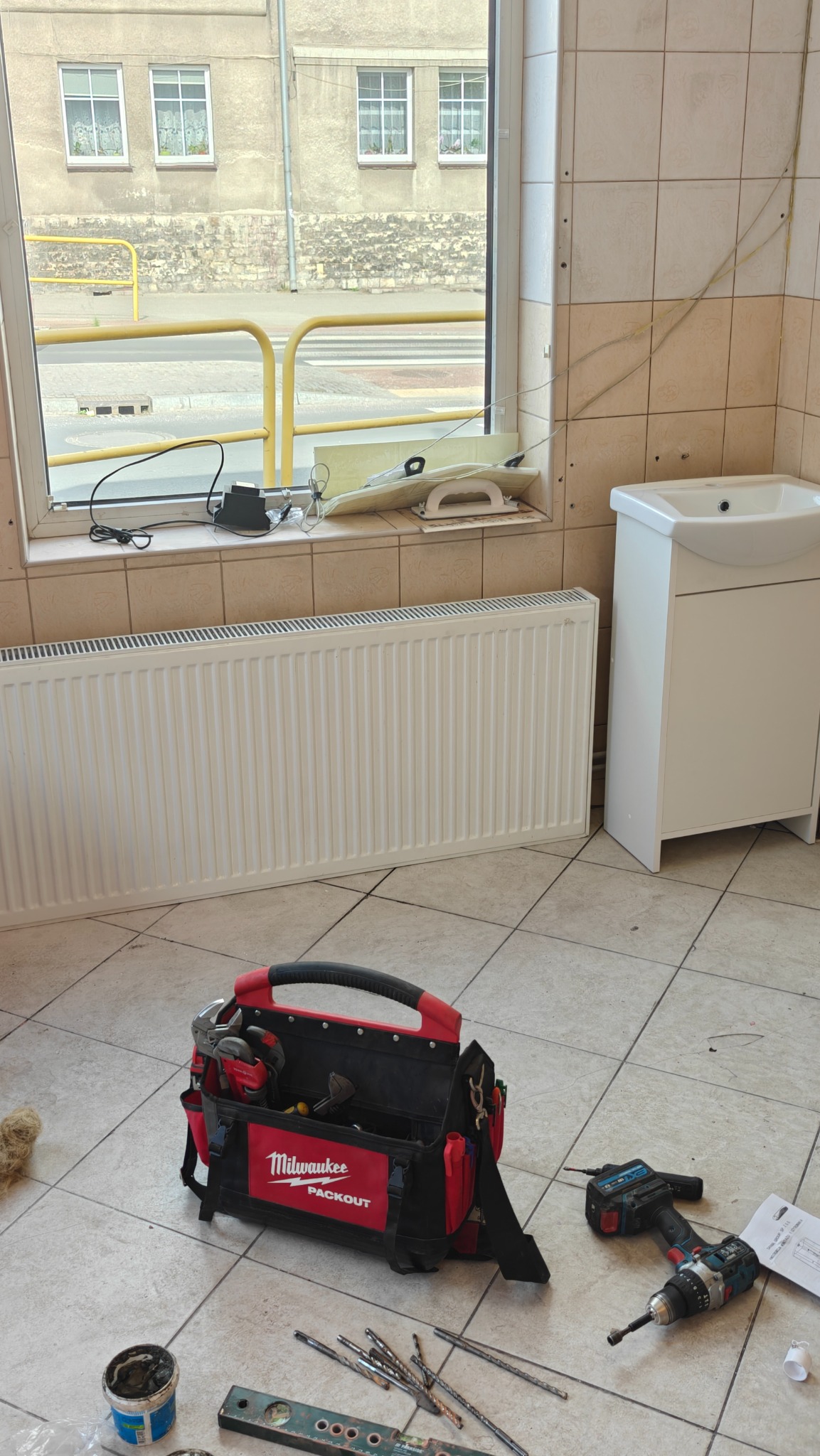 Biały grzejnik panelowy pod oknem w łazience z beżowymi płytkami. Na podłodze torba narzędziowa Milwaukee, wiertarka i poziomica. Widok na ulicę przez okno.