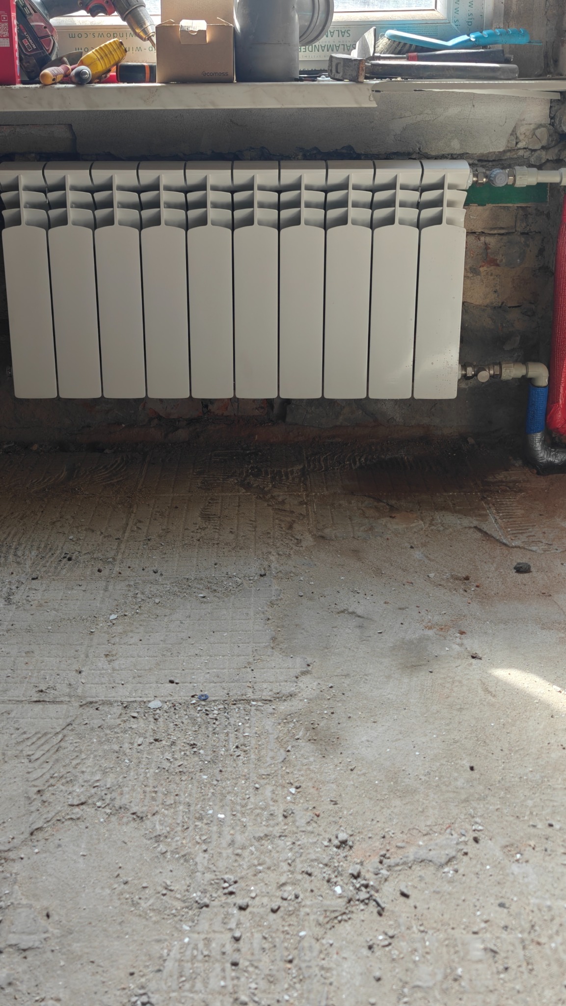 Biały, panelowy grzejnik zamontowany na ścianie z widocznymi śladami prac instalacyjnych. Na parapecie narzędzia i materiały budowlane. Surowa podłoga w tle.
