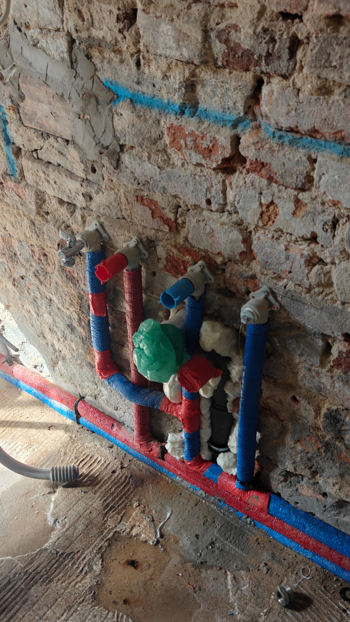 Instalacja wodna z rurami w kolorach czerwonym i niebieskim, zabezpieczona pianką montażową, na tle surowej ściany z cegły i betonu. Widoczne zawory i fragment podłogi.