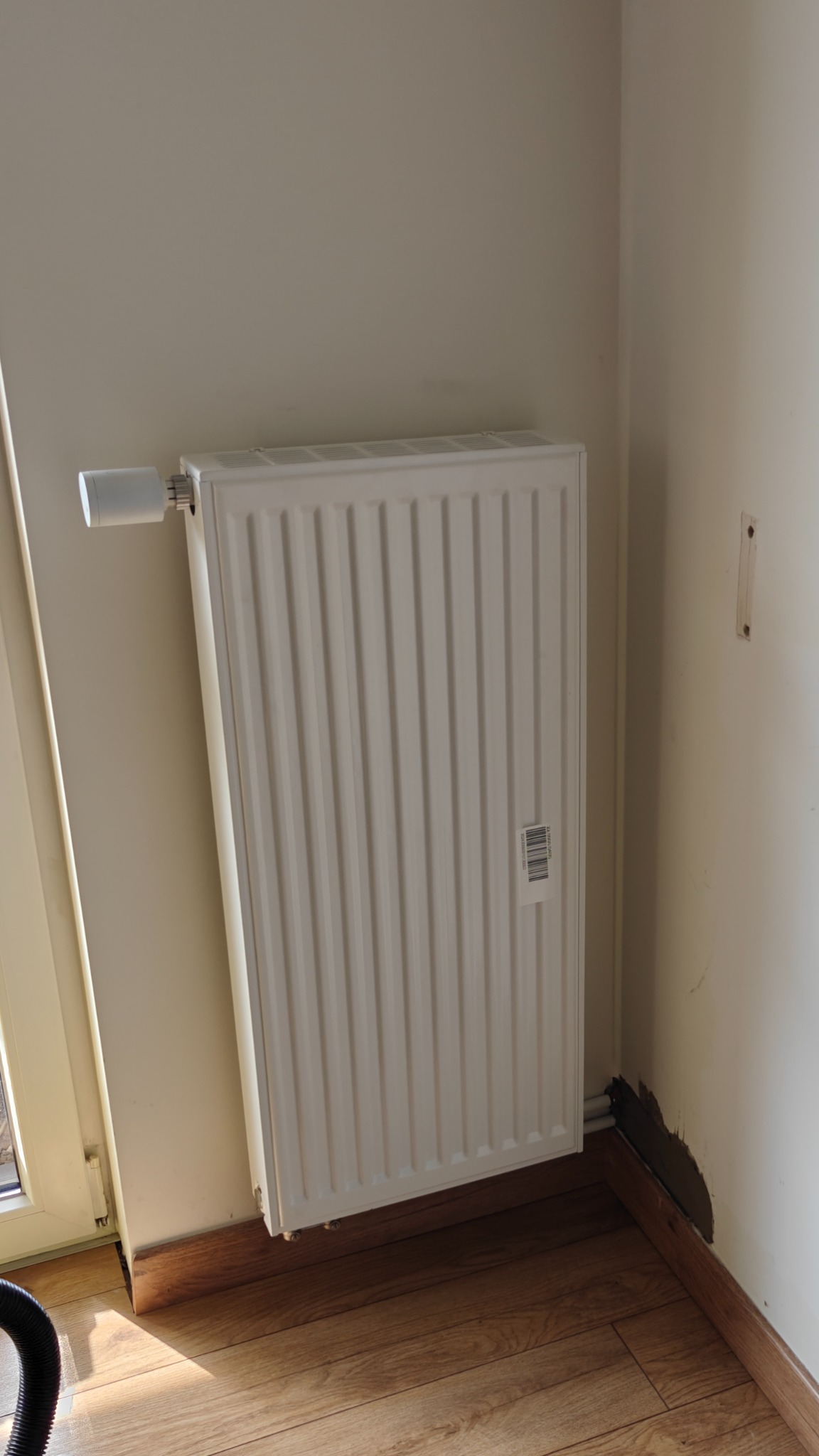Biały grzejnik panelowy z termostatem, zamontowany przy ścianie w rogu pokoju z drewnianą podłogą. Widoczne ślady po remoncie przy listwie przypodłogowej.