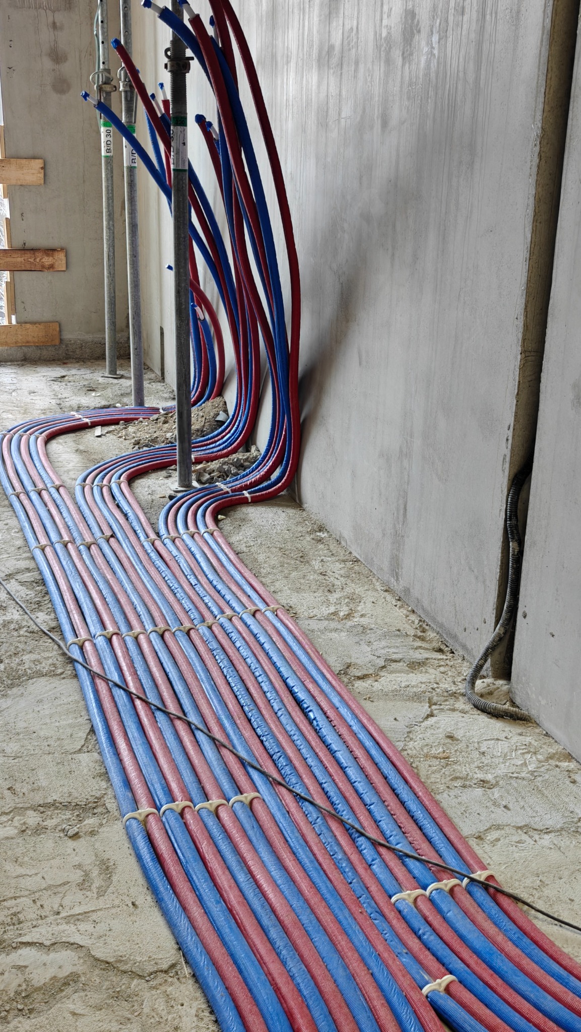 Równolegle ułożone, czerwono-niebieskie rury instalacji grzewczej na podłożu w surowym wnętrzu, zabezpieczone uchwytami, prowadzone wzdłuż ściany z betonu.