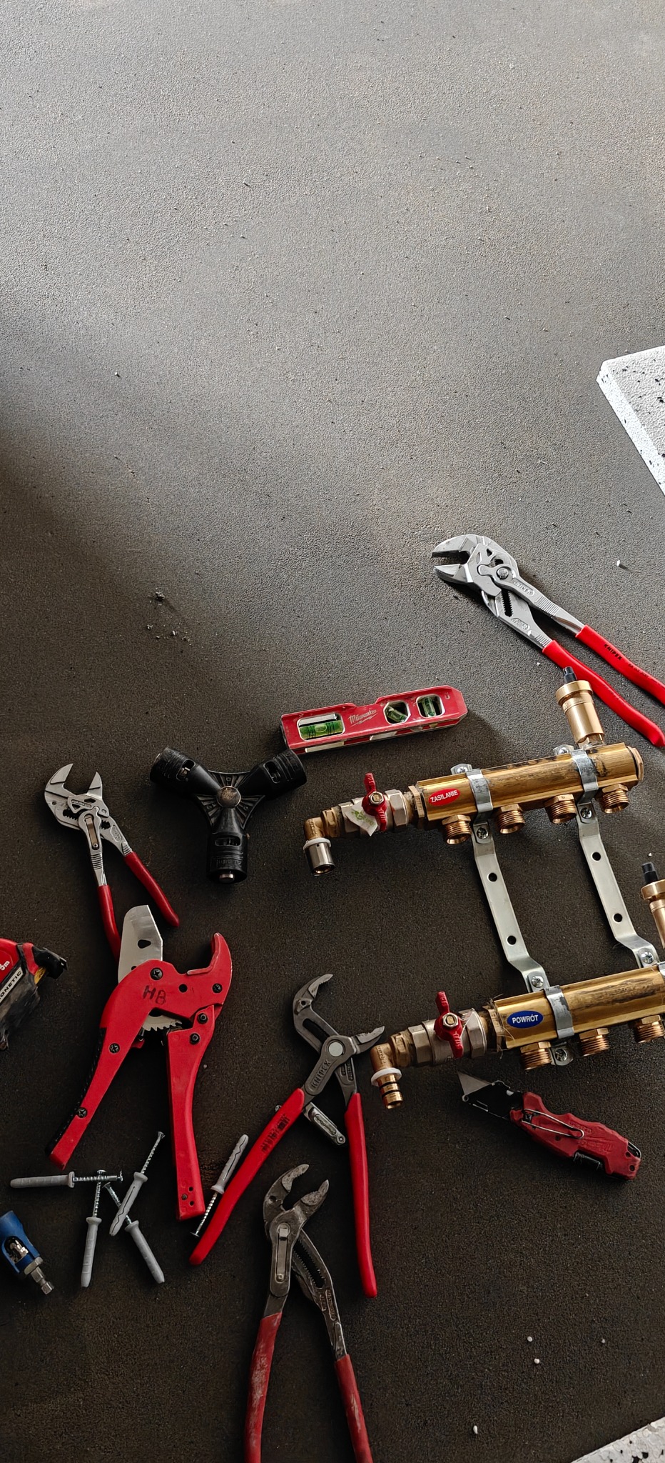 Rozdzielacz mosiężny z zaworami, poziomica i zestaw narzędzi (klucze, nożyce do rur) na szarym tle. Widok z góry, ostre światło.