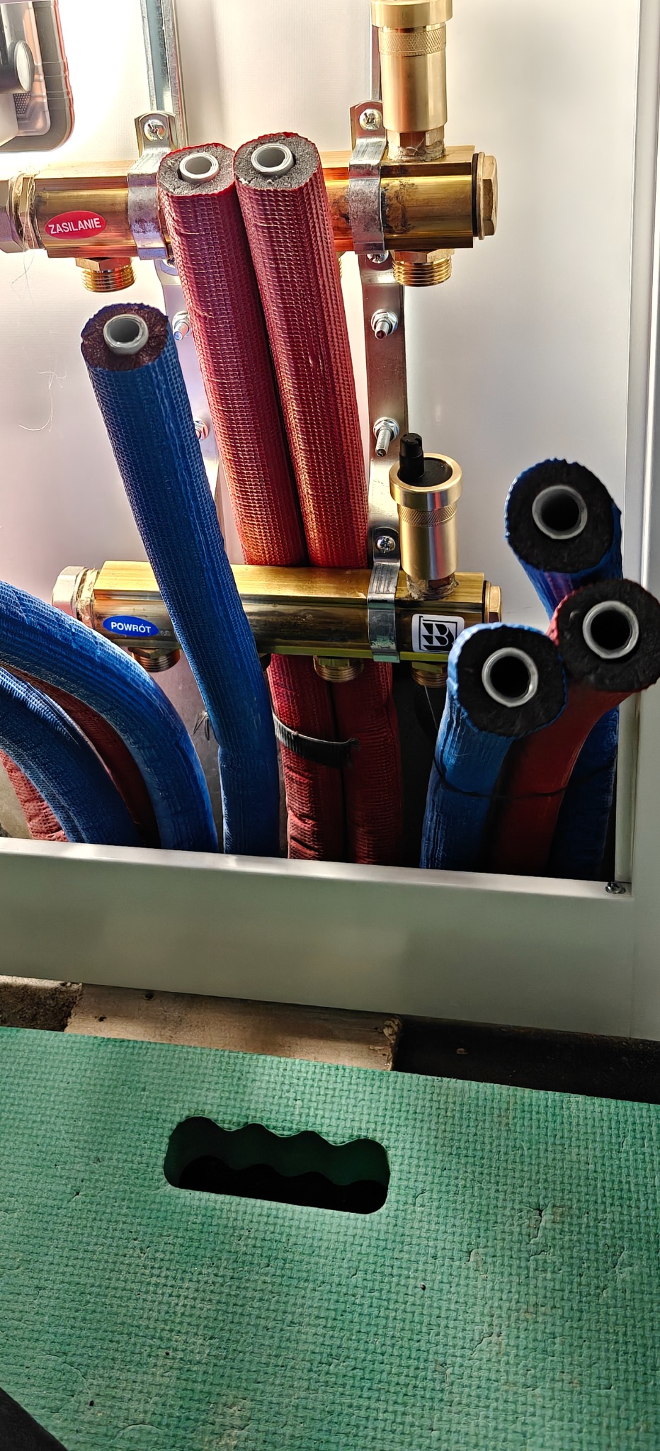 Rozdzielacz instalacji grzewczej z rurami w izolacji (czerwone - zasilanie, niebieskie - powrót) zamontowany w szafce. Widoczne elementy mosiężne i stalowe.