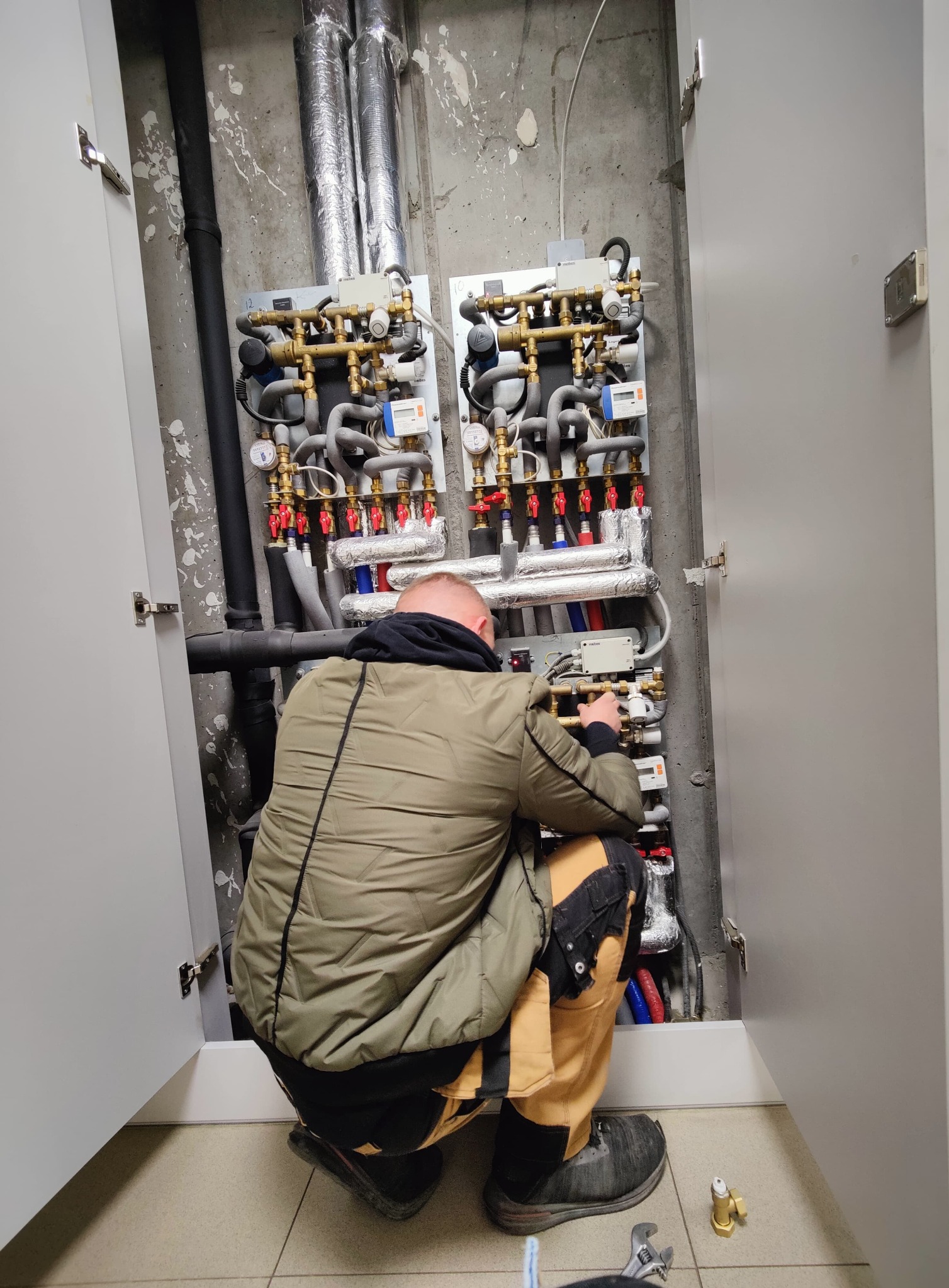 Hydraulik w trakcie montażu rozdzielacza instalacji grzewczej w szafce. Widoczne rury, zawory i manometry. Na podłodze leżą narzędzia.