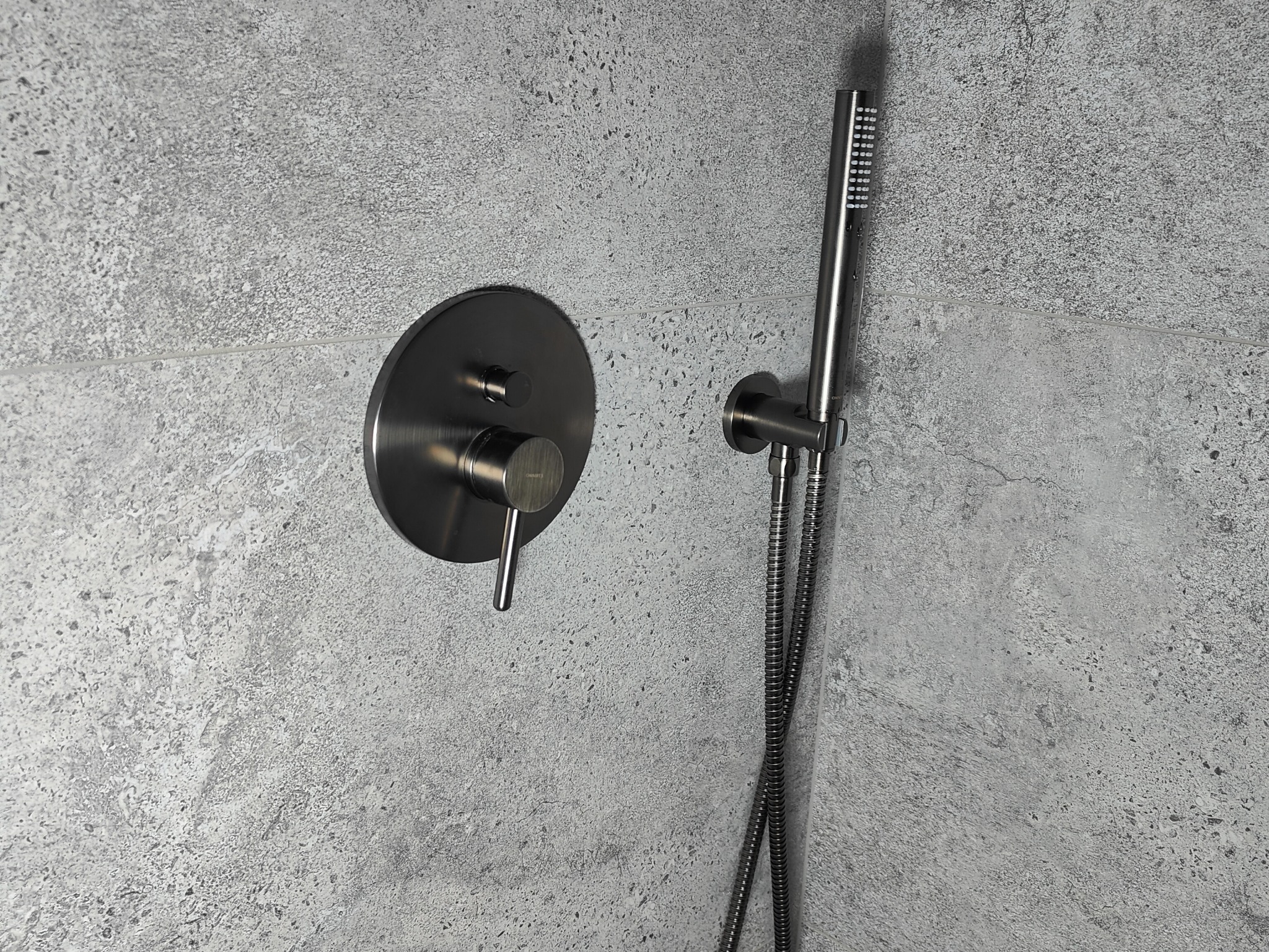 Nowoczesna, metalowa bateria prysznicowa w kolorze grafitowym, zamontowana na szarej, teksturowanej ścianie. Minimalistyczny design, widoczny wąż i słuchawka prysznicowa.