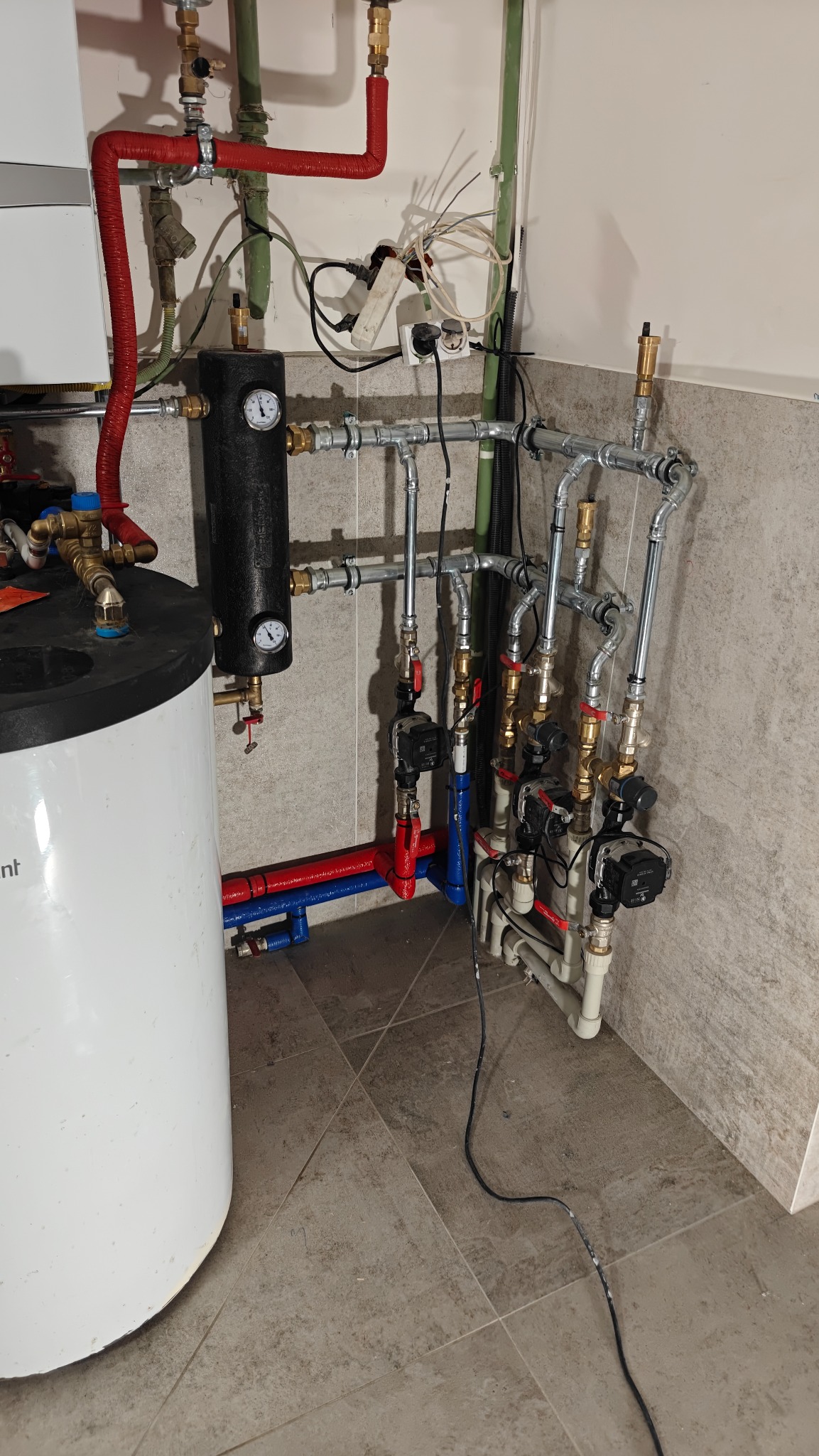 Złożona instalacja grzewcza z kotłem, rurami miedzianymi i zaworami, widoczna na tle szarej ściany i podłogi z płytek. Elementy instalacji w kolorach czerwonym, niebieskim i srebrnym.