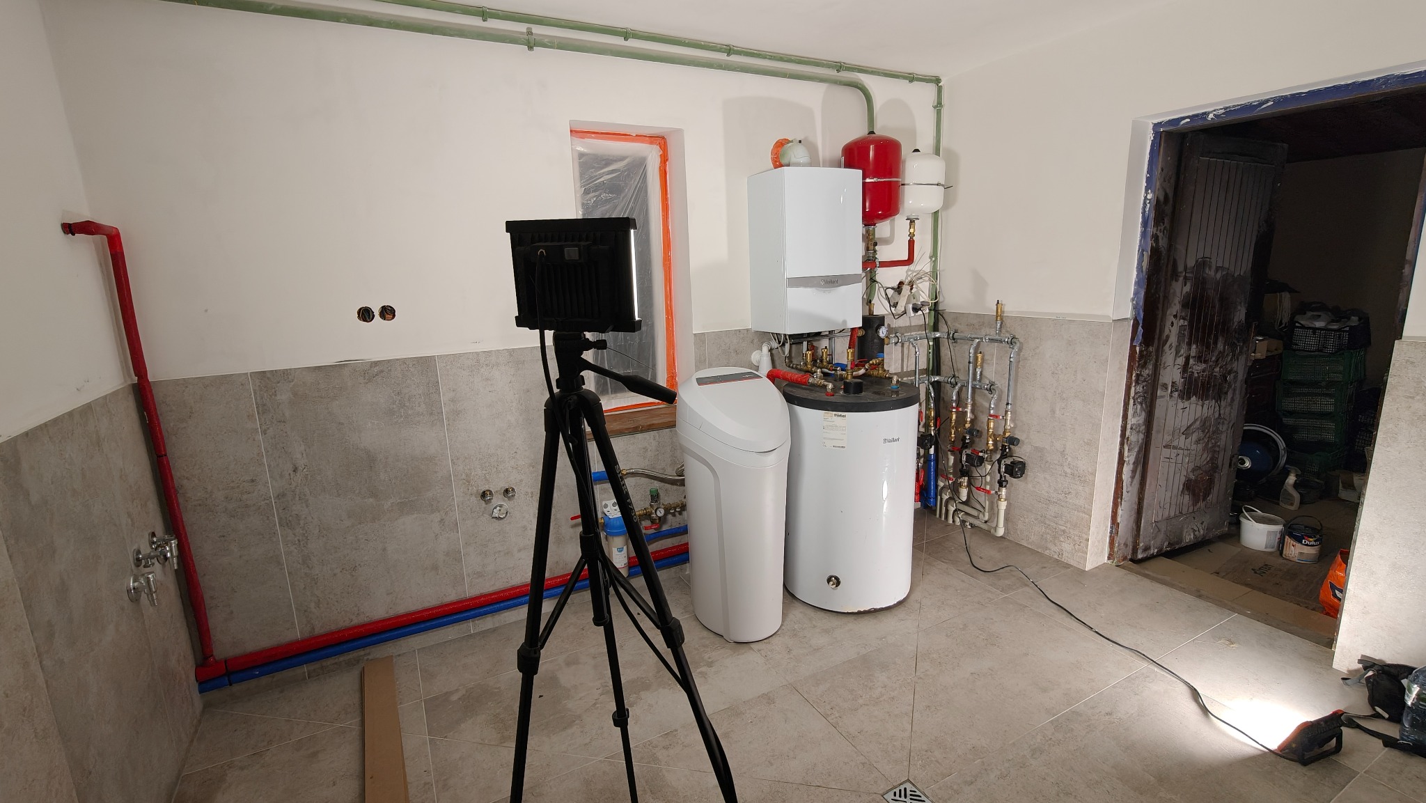 Nowoczesny piec gazowy Vaillant z zasobnikiem i zmiękczaczem wody, oświetlony lampą na statywie, na tle wykafelkowanej ściany i instalacji rurowej.