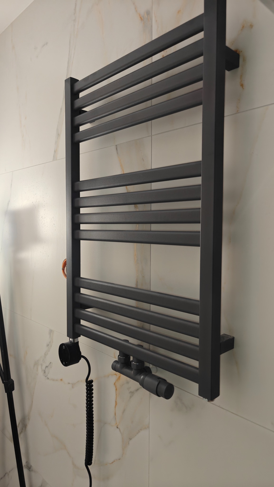 Grafitowy grzejnik łazienkowy drabinkowy, zamontowany na ścianie z marmurowym wzorem. Widoczne zawory termostatyczne i element grzejny z przewodem.