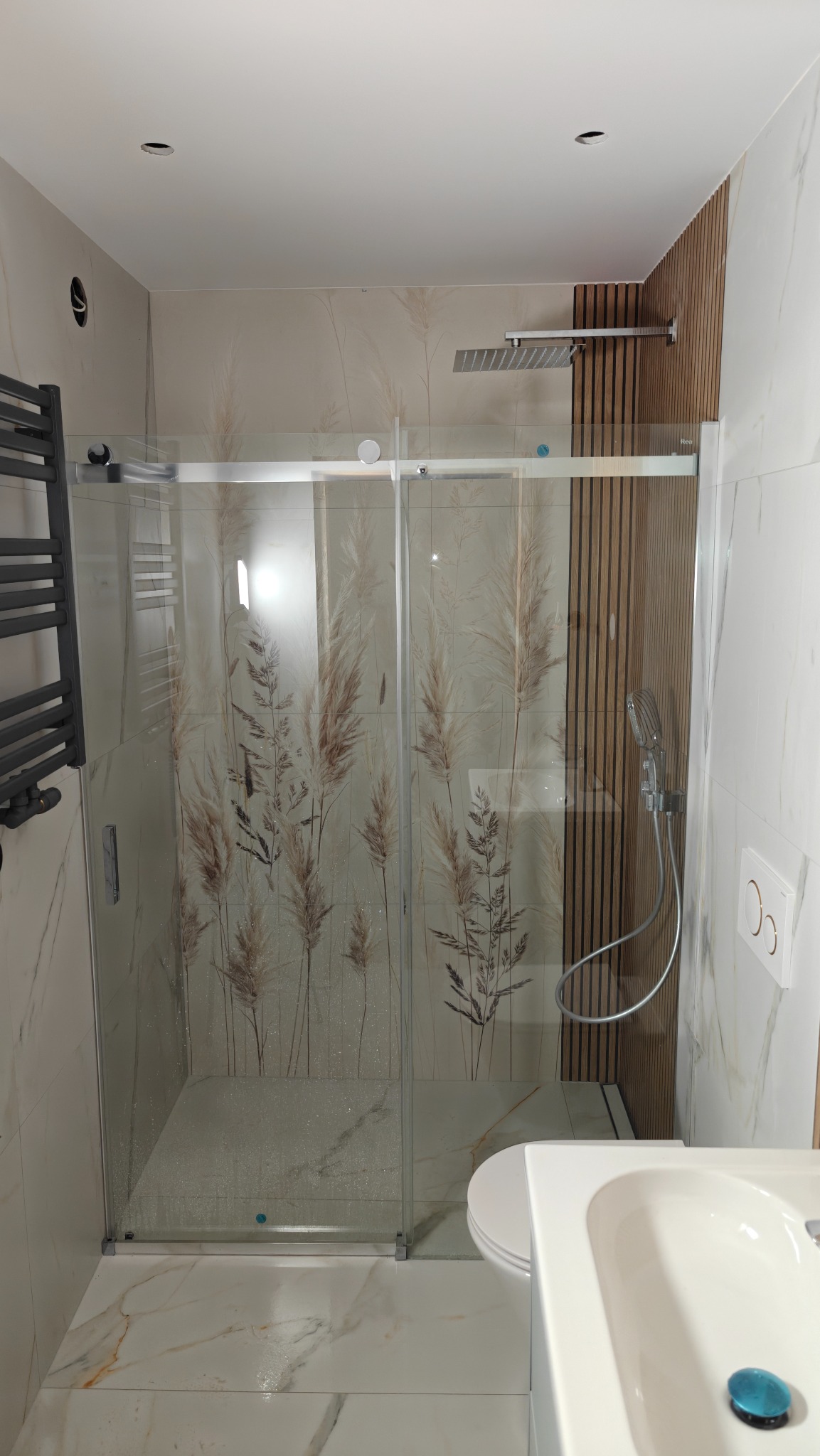 Nowoczesna kabina prysznicowa z motywem roślinnym na szklanych drzwiach, w jasnej łazience z marmurowymi płytkami i drewnianymi panelami dekoracyjnymi.