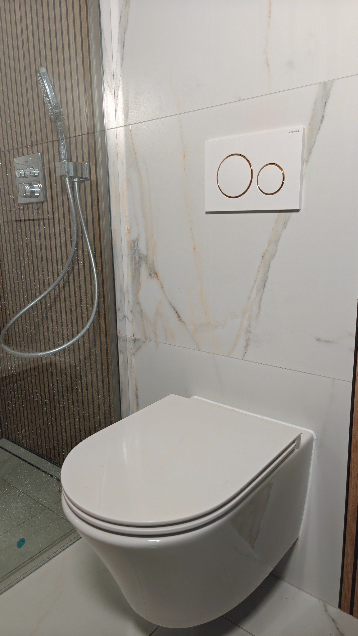 Nowoczesna toaleta podwieszana z przyciskiem spłukującym na marmurowej ścianie, obok kabiny prysznicowej w Świętochłowicach. Minimalistyczny design.