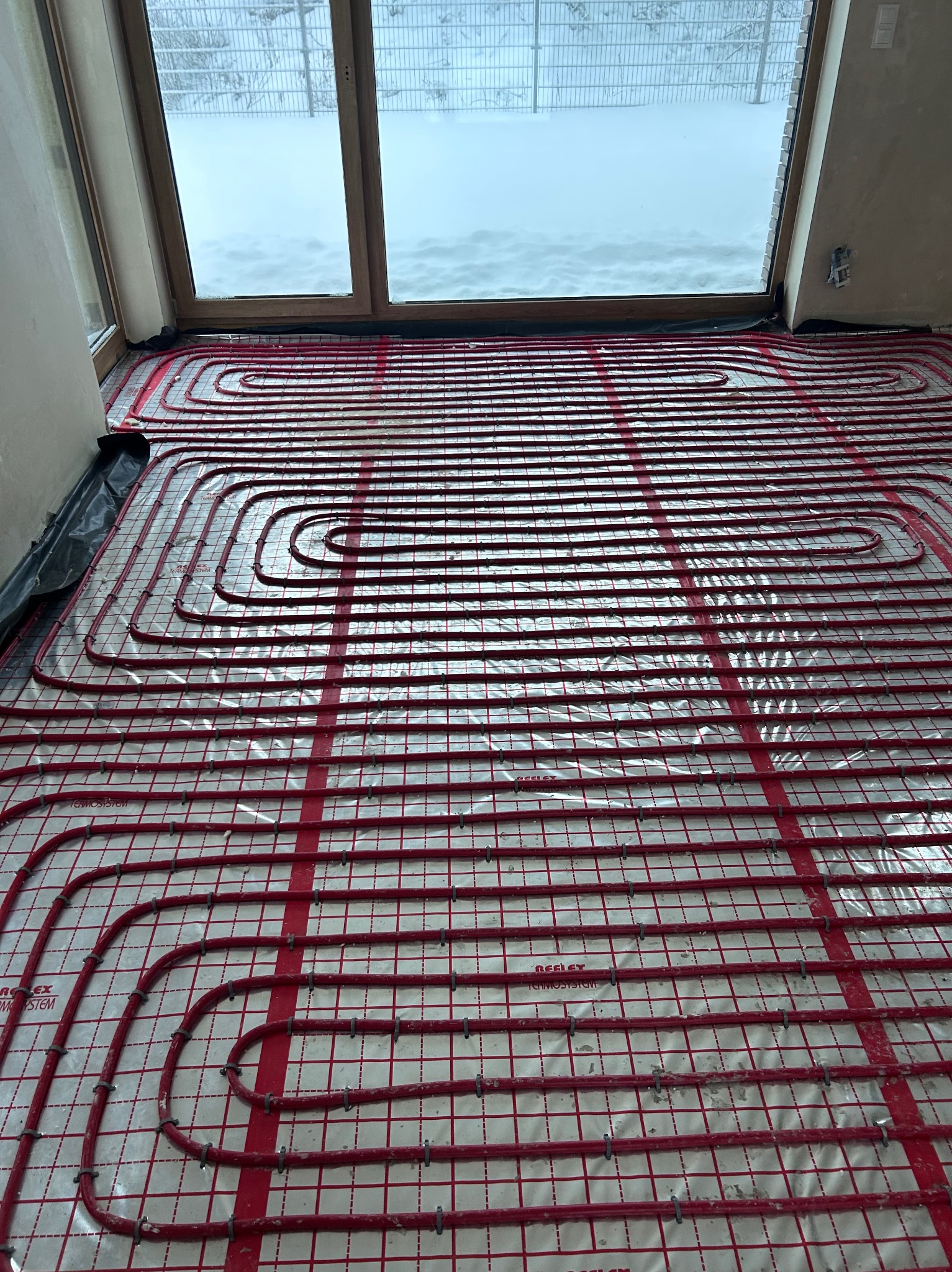Czerwone rury ogrzewania podłogowego ułożone na folii z siatką, widok z góry, okno z widokiem na zaśnieżony ogród w tle. Etap instalacji przed wylewką.