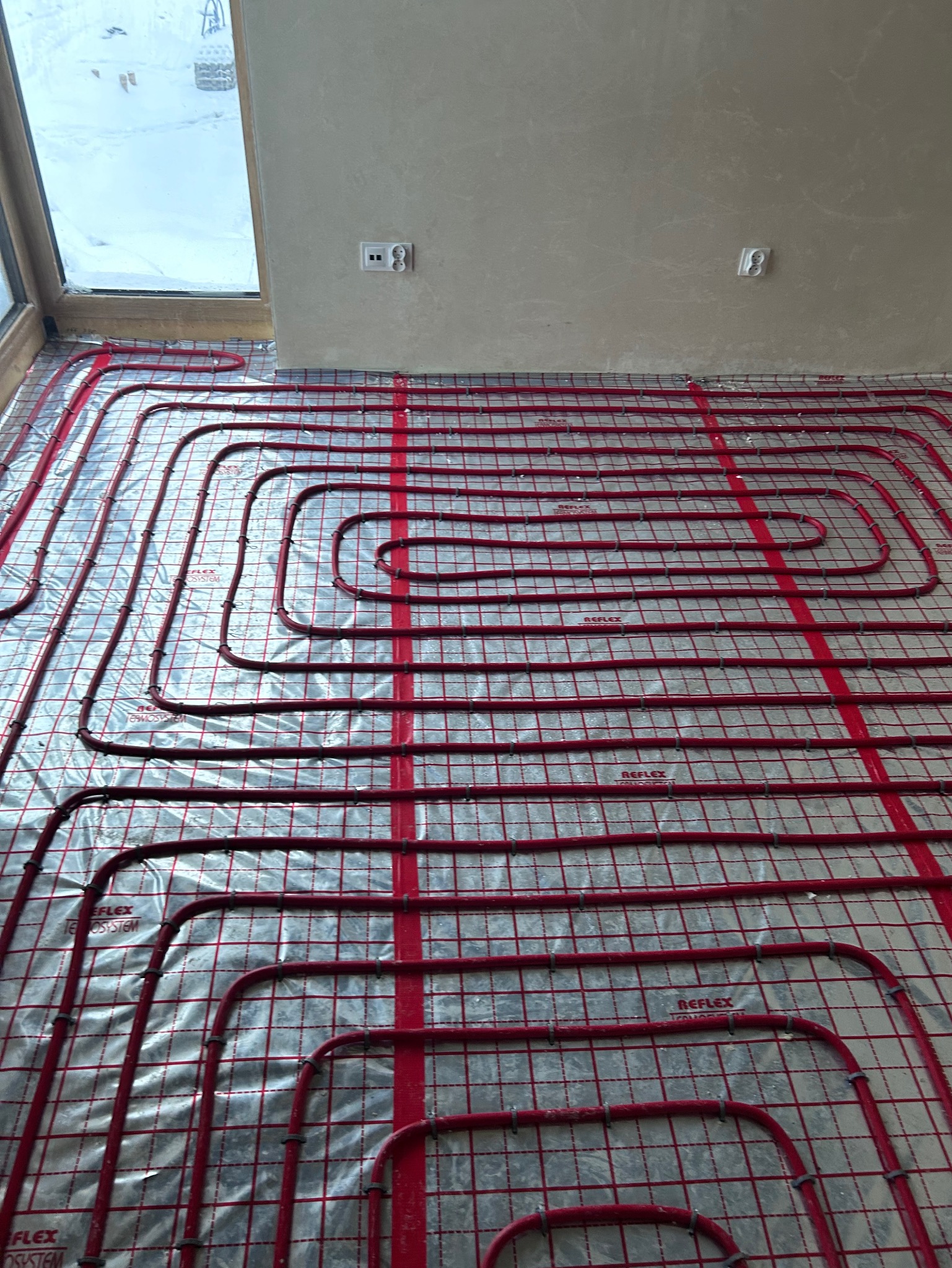 Czerwone rury ogrzewania podłogowego ułożone na srebrnej folii z siatką, widoczne gniazdka elektryczne na ścianie i okno w tle. System Reflex.