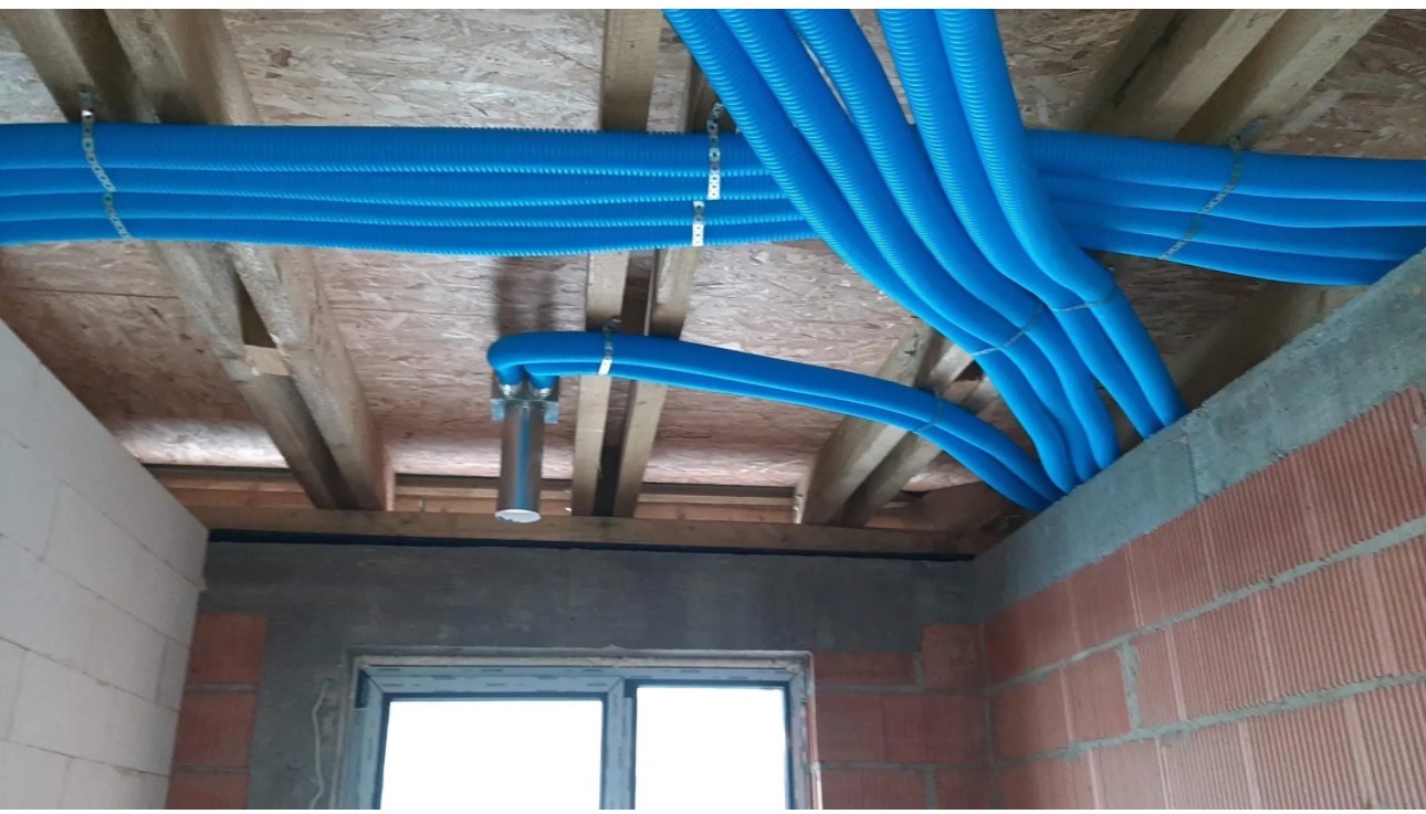 Niebieskie rury instalacyjne, przymocowane do drewnianego stropu, widoczne w surowym wnętrzu budynku z oknem i ścianami z cegły. Instalacja w trakcie realizacji.