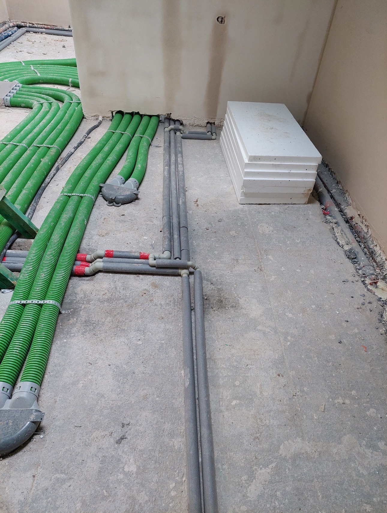Wnętrze w trakcie prac: zielone rury karbowane i szare rury instalacyjne ułożone na podłodze, obok stos białych płyt izolacyjnych. Widoczne fragmenty ścian.