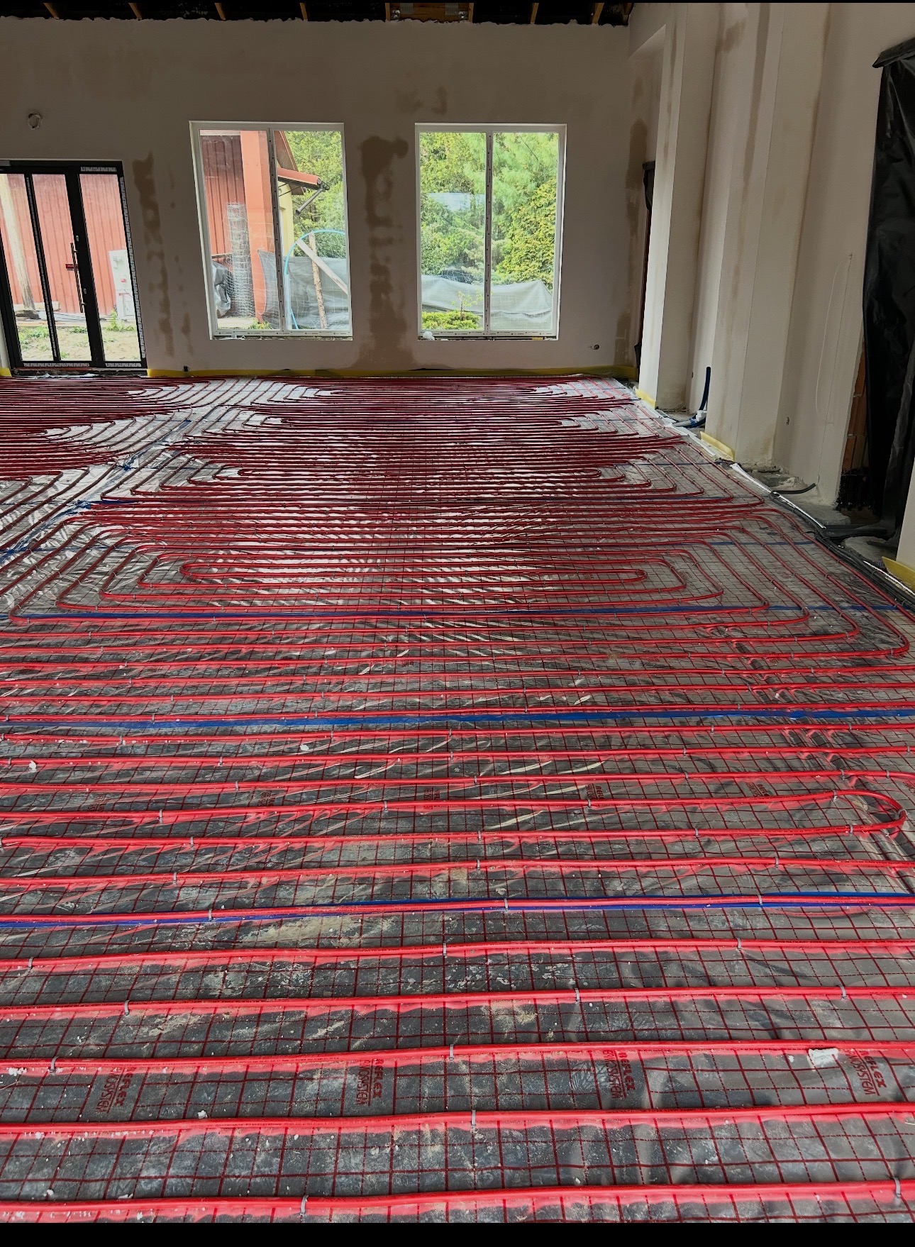 Czerwone rury ogrzewania podłogowego ułożone na czarnej macie w jasnym pomieszczeniu z oknami. Widoczne fragmenty ścian i przygotowania do dalszych prac wykończeniowych.