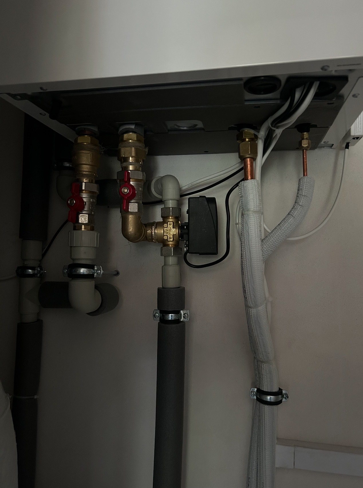 Szczegółowe ujęcie instalacji hydraulicznej z zaworami, rurami izolowanymi i przewodami, zamontowanej pod urządzeniem. Widoczne elementy z mosiądzu i tworzywa.