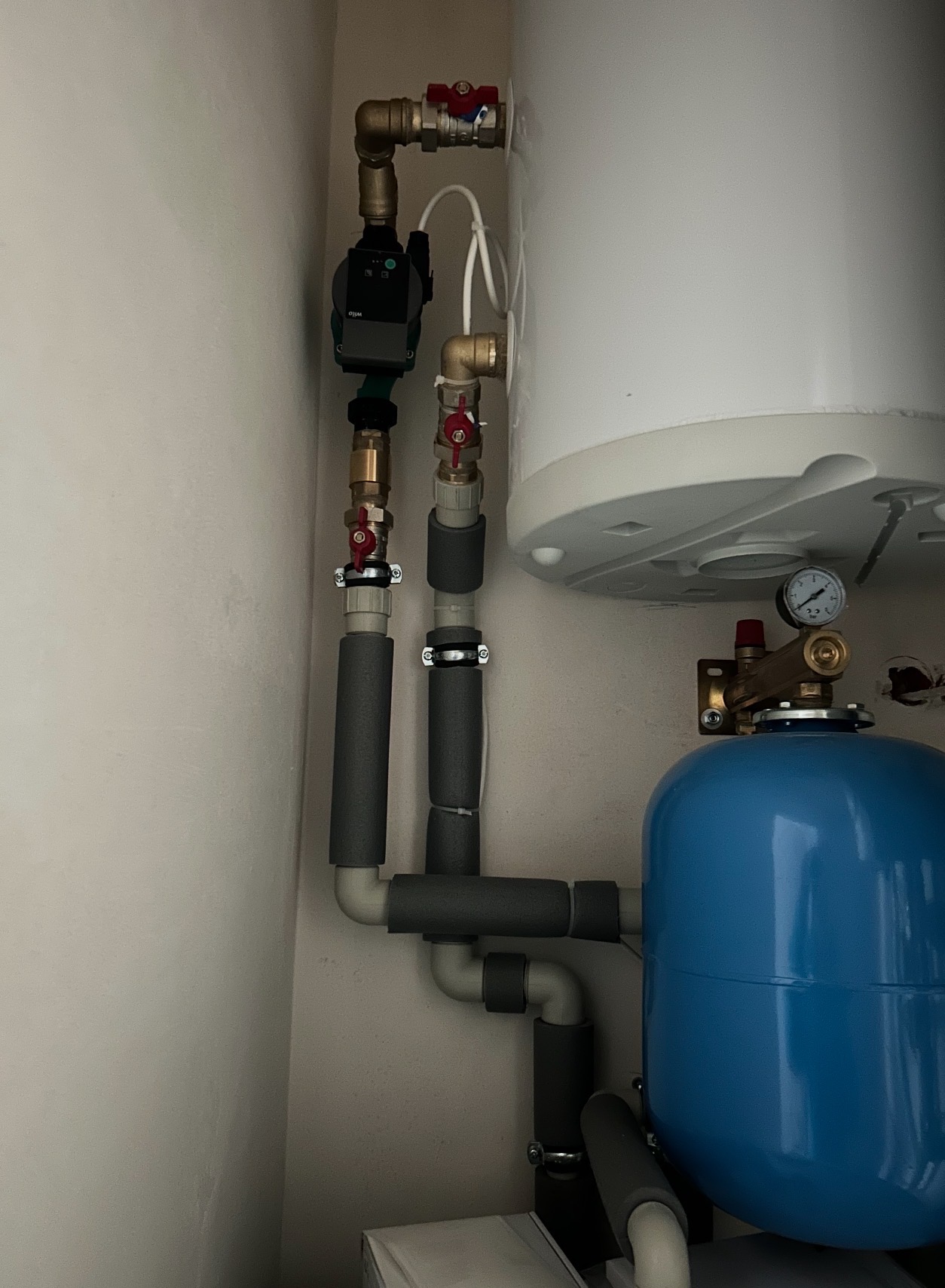 Zamontowany biały bojler z niebieskim zbiornikiem wyrównawczym, widoczne rury miedziane i szare izolowane, zawory z czerwonymi kurkami, manometr.