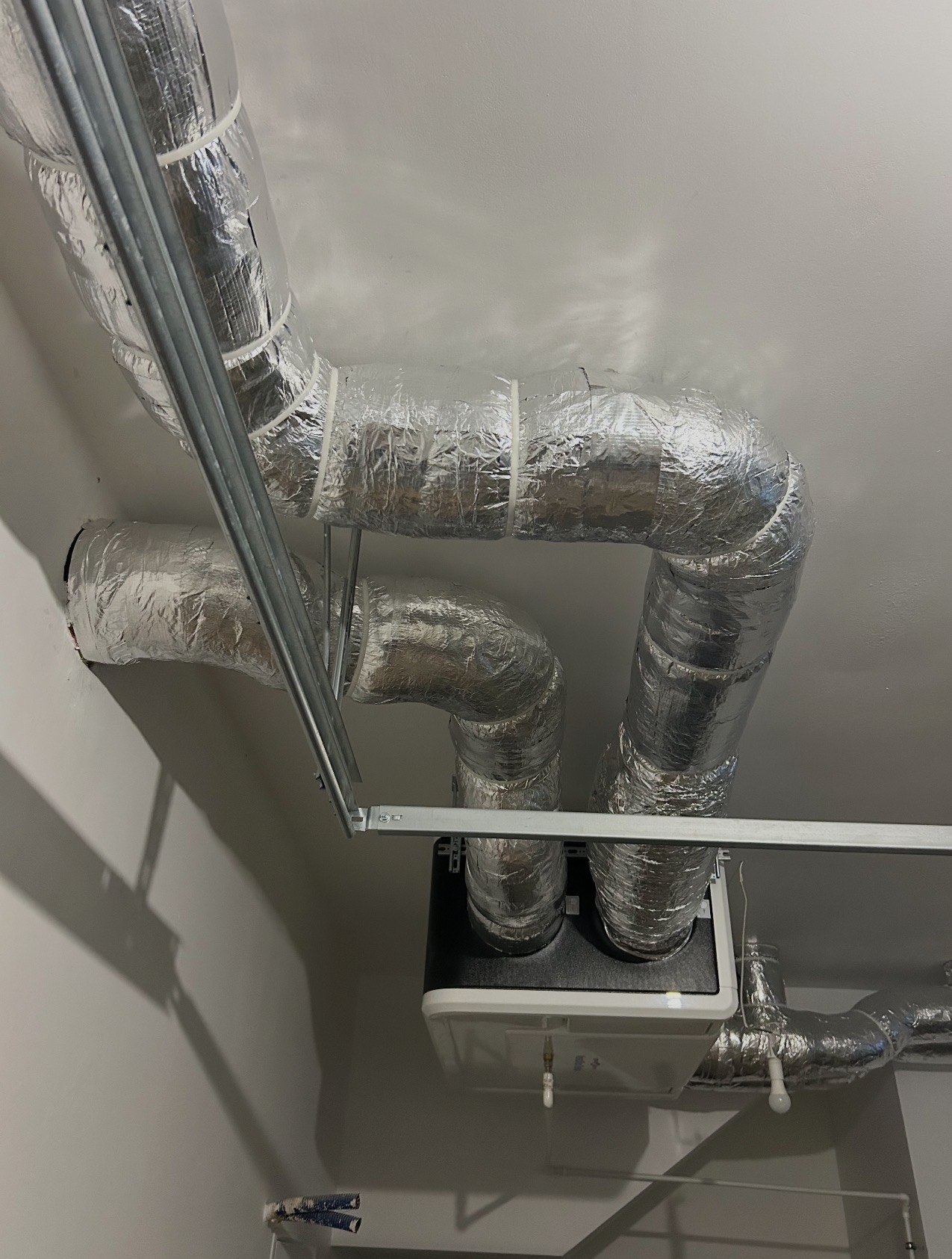 Izolowane przewody wentylacyjne z centralą rekuperacyjną zamontowane pod sufitem, widoczne elementy mocujące i fragment ściany. Srebrna izolacja na rurach.