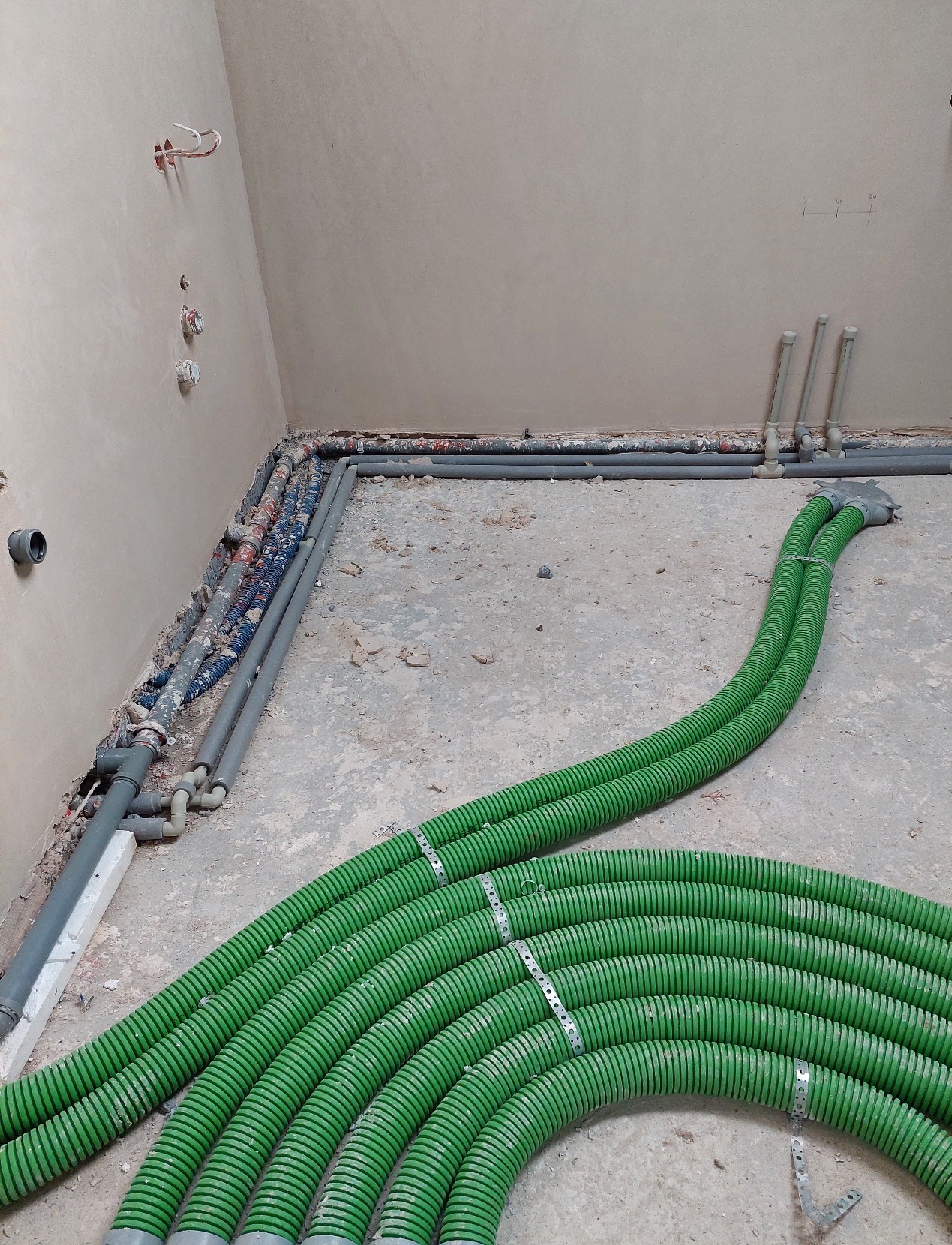 Zielone rury instalacji grzewczej ułożone na podłodze w surowym wnętrzu. Widoczne także szare rury i instalacje w ścianie. Etap prac instalacyjnych.