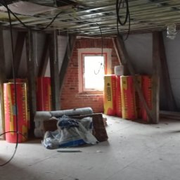 MM-BUD - Poddasze w trakcie ocieplania: widoczne materiały izolacyjne Rotaflex, konstrukcja sufitu podwieszanego i okna w szpalecie z cegły. Prace wykończeniowe w toku.