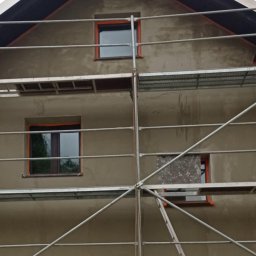 MM-BUD - Dom w trakcie tynkowania elewacji, widoczne rusztowania i okna z pomarańczowymi ramami. Częściowo otynkowana ściana szczytowa. Prace w toku.