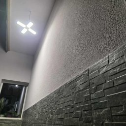 MM-BUD - Ściana z dekoracyjną, ciemną okładziną kamienną w dolnej części i jasnym tynkiem strukturalnym powyżej. Widoczne okno z roślinami i nowoczesna lampa sufitowa.