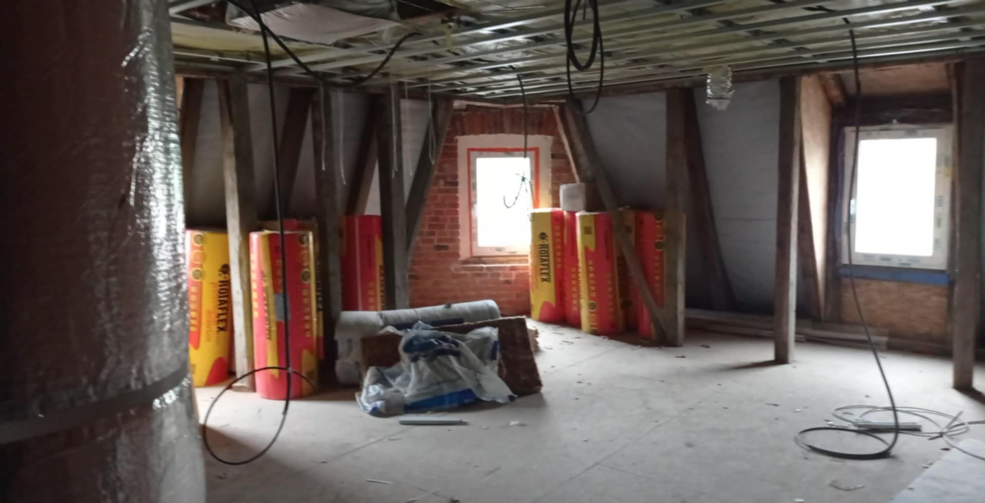 Poddasze w trakcie ocieplania: widoczne materiały izolacyjne Rotaflex, konstrukcja sufitu podwieszanego i okna w szpalecie z cegły. Prace wykończeniowe w toku.