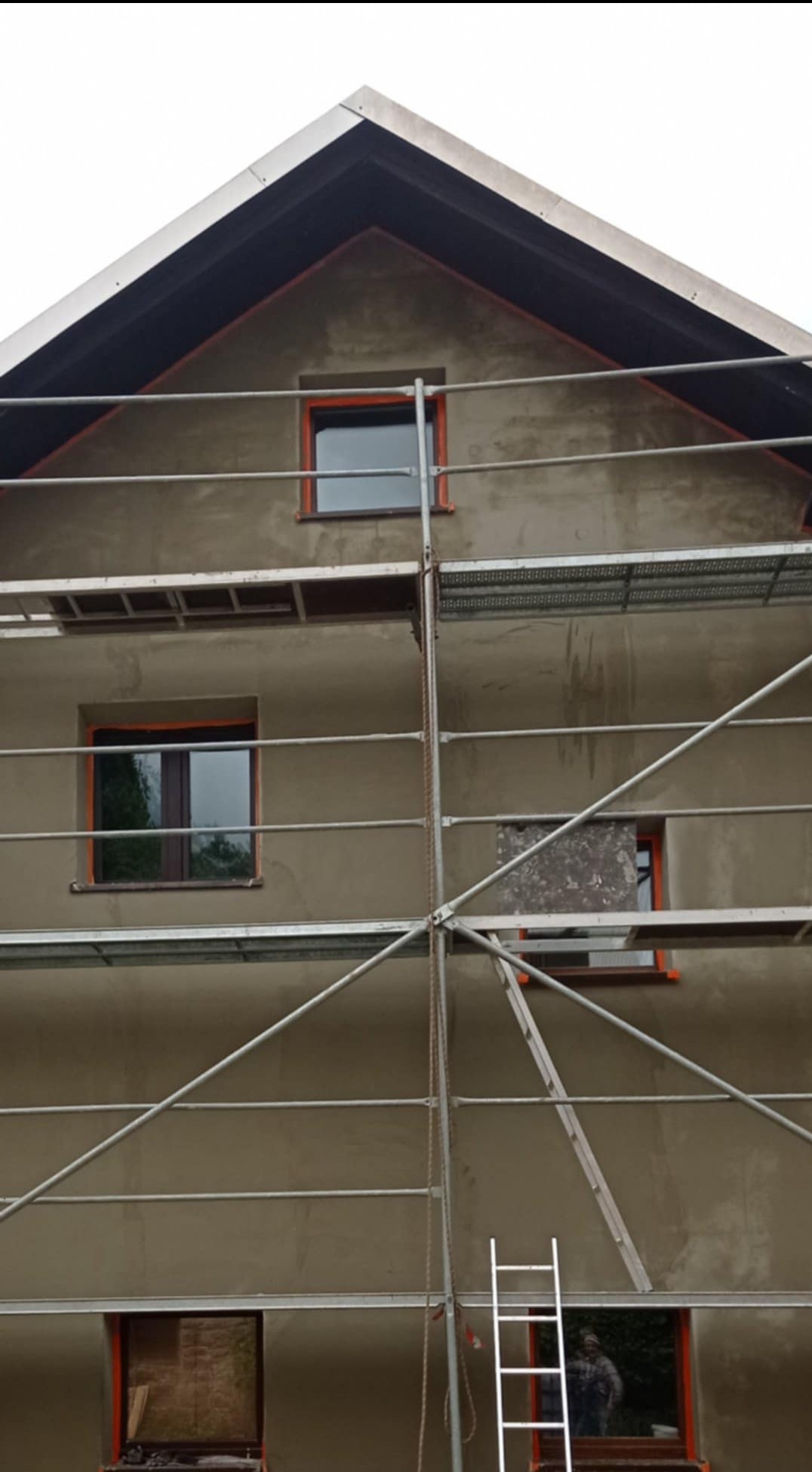 Dom w trakcie tynkowania elewacji, widoczne rusztowania i okna z pomarańczowymi ramami. Częściowo otynkowana ściana szczytowa. Prace w toku.