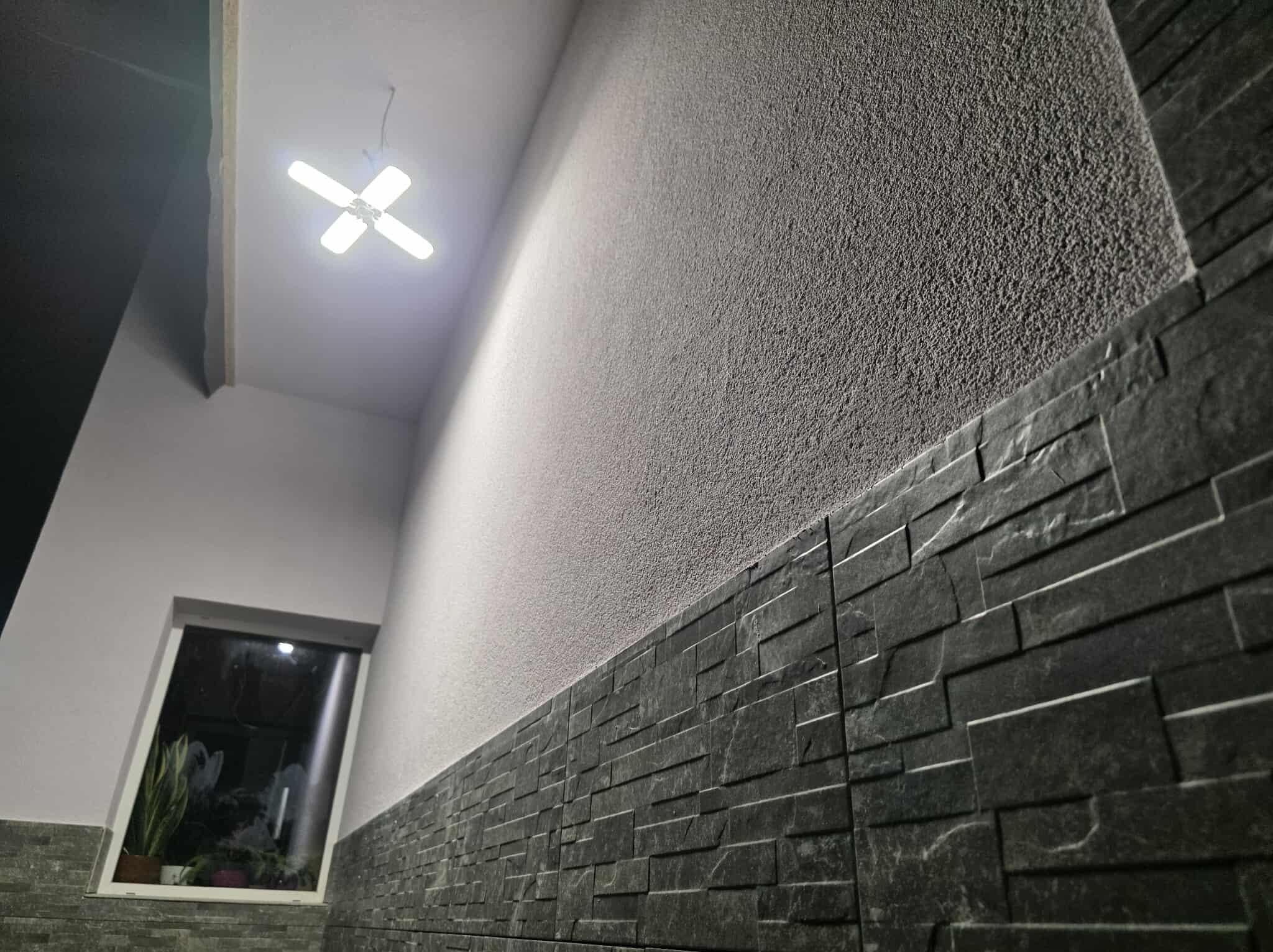 Ściana z dekoracyjną, ciemną okładziną kamienną w dolnej części i jasnym tynkiem strukturalnym powyżej. Widoczne okno z roślinami i nowoczesna lampa sufitowa.