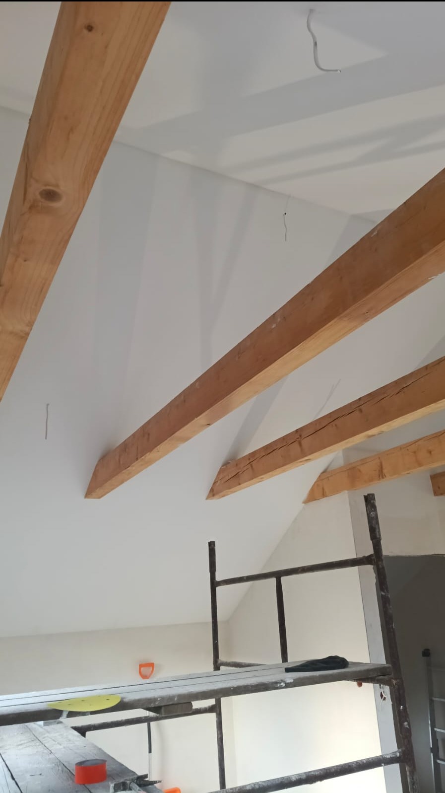 Białe wnętrze poddasza z drewnianymi belkami stropowymi, widok z rusztowania. Świeżo pomalowane ściany i sufit, widoczne kable. Ujęcie z dołu, perspektywa z rusztowania.