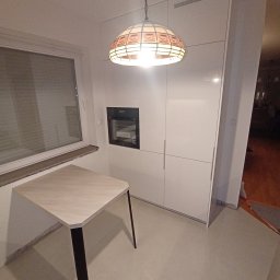 Meble Korczak - Biała zabudowa kuchenna z piekarnikiem w ścianie, oświetlona lampą w stylu Tiffany. Blat stołu przylega do okna. Minimalistyczny design.