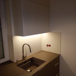 Meble Korczak - Nowoczesna zabudowa kuchenna z szarymi frontami, podświetleniem LED i stalową baterią. Widoczne gniazdka elektryczne na ścianie i okno z żaluzjami.