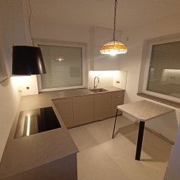 Meble Korczak - Jasna kuchnia na wymiar z beżowymi frontami, blatem w kolorze betonu i lampą w stylu Tiffany. Widok na okno z żaluzjami i stół z czarną nogą.