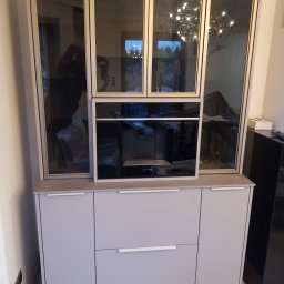 Meble Korczak - Jasnoszara zabudowa kuchenna z przeszklonymi szafkami wiszącymi i wbudowanym ekspresem do kawy Siemens. Minimalistyczny design, nowoczesne oświetlenie w tle.