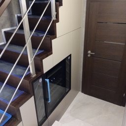 Meble Korczak - Schody z zabudową meblową i wbudowaną chłodziarką na wino, brązowe stopnie zabezpieczone folią, białe fronty, drzwi wewnętrzne w kolorze wenge.