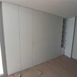 Meble Korczak - Długa, biała szafa wnękowa z otwartymi drzwiami ukazującymi półki. Podłoga zabezpieczona kartonem. Minimalistyczny design, jasne wnętrze.