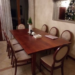 Meble Korczak - Drewniany stół z krzesłami w jadalni, nakryty do świątecznego posiłku. W tle okno z firankami i lustro z odbiciem choinki. Na podłodze wzór zegara słonecznego.