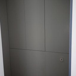 Meble Korczak - Szara, minimalistyczna szafa wnękowa z płaskimi frontami, z widocznym czujnikiem dymu. Proste linie i stonowana kolorystyka.