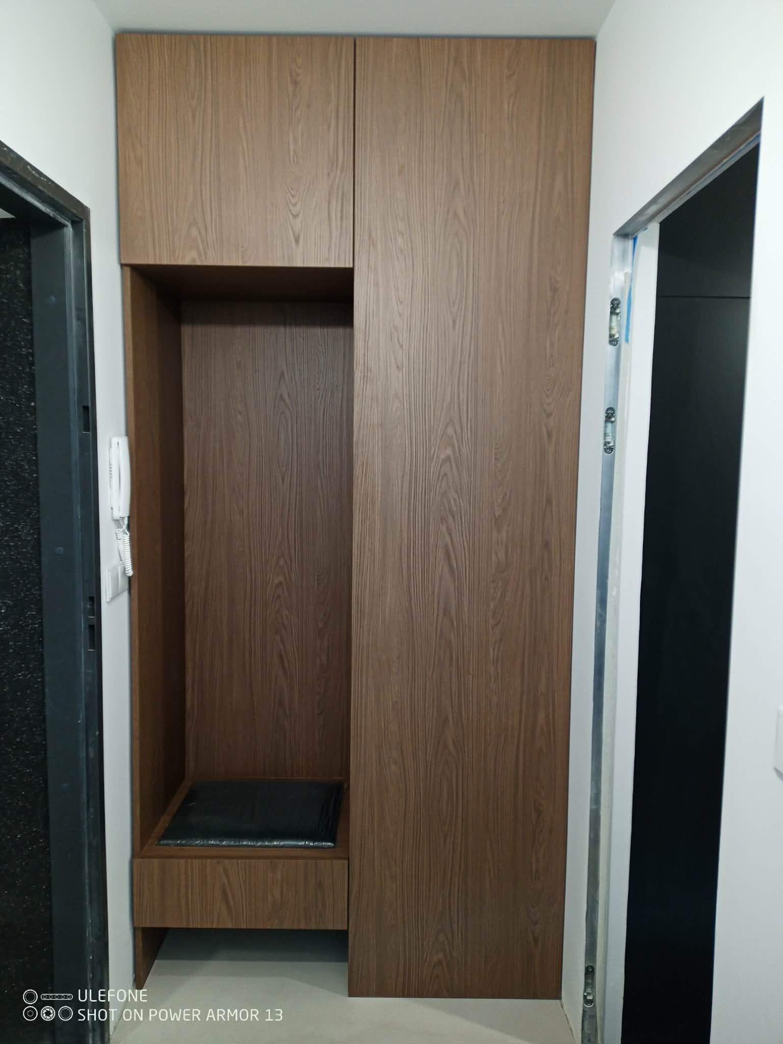 Funkcjonalna szafa w przedpokoju z siedziskiem i półką, wykończona okleiną w odcieniach brązu. Widoczny domofon po lewej stronie i fragment otwartych drzwi po prawej.