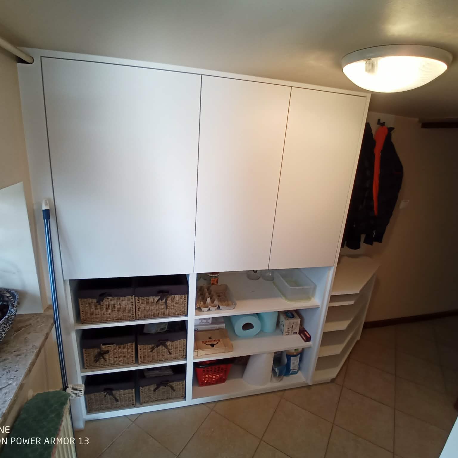 Biała szafa wnękowa z półkami i koszami, widok z boku, oświetlenie sufitowe, płytki podłogowe w odcieniach beżu. Widoczne przedmioty na półkach.