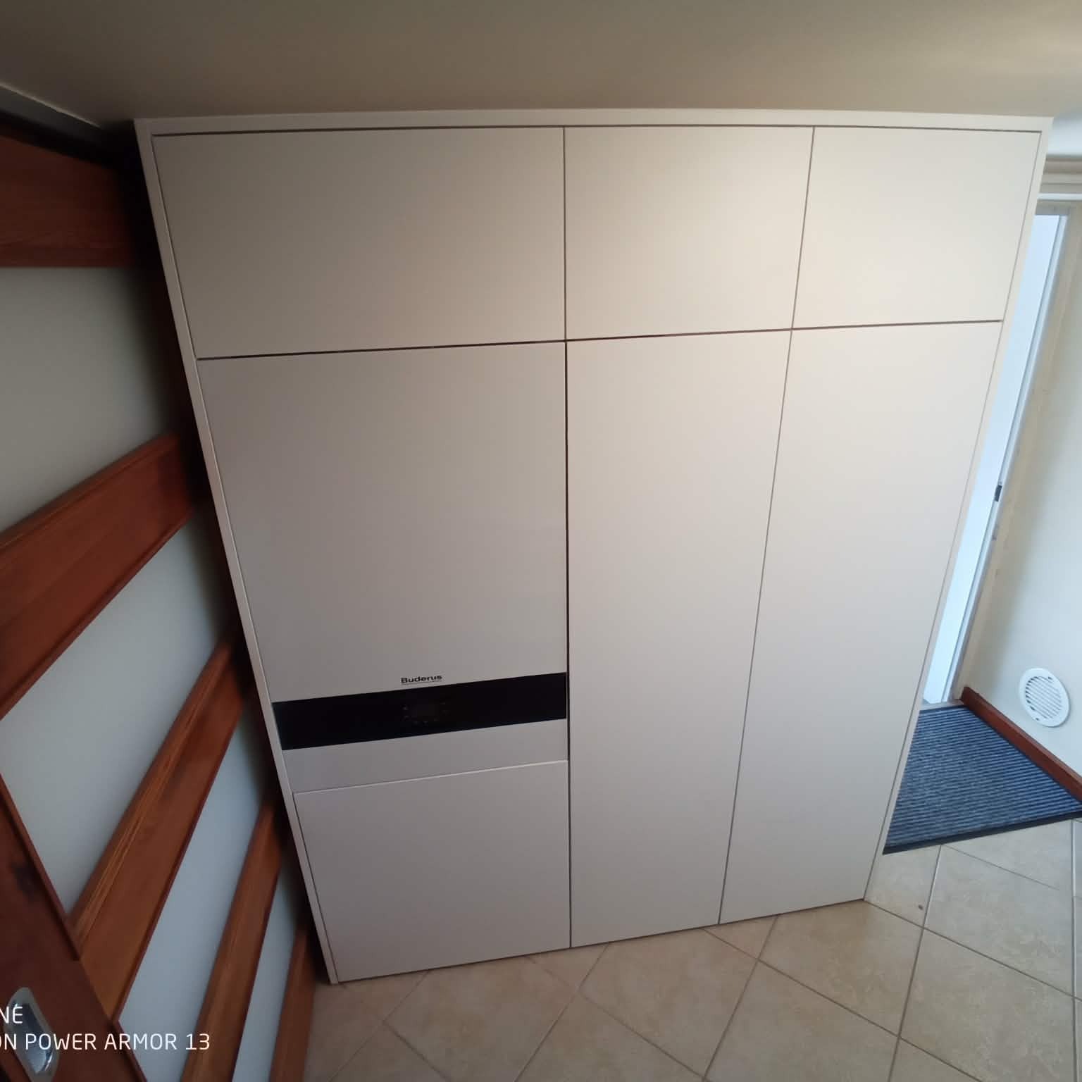 Biała szafa wnękowa z zabudowanym piecem gazowym Buderus, widoczna z perspektywy podłogi, z elementami drewnianej framugi drzwi po lewej stronie.