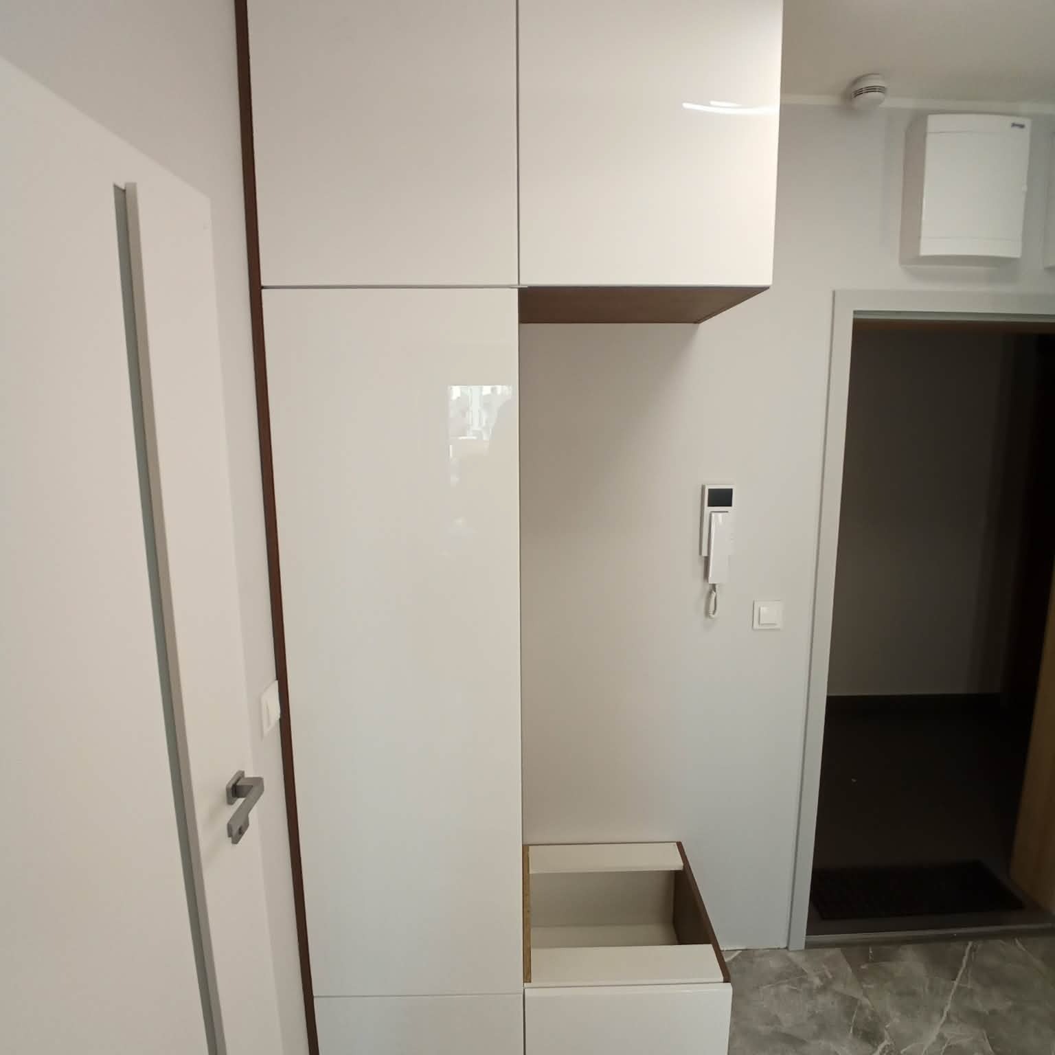 Biała, lakierowana zabudowa meblowa w przedpokoju z siedziskiem i szafkami. Minimalistyczny design, szare płytki podłogowe, widoczny domofon i drzwi.
