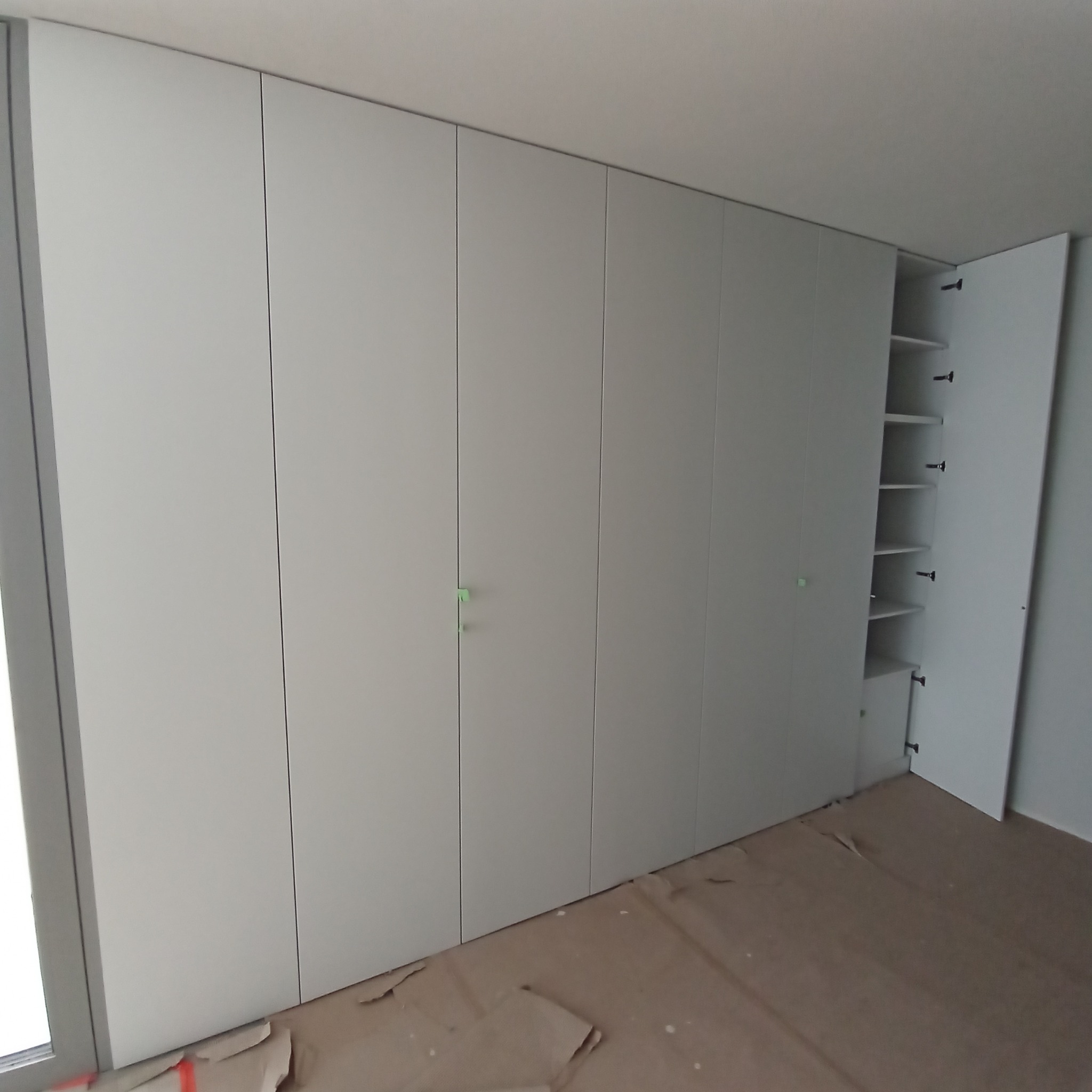 Biała szafa wnękowa z półkami, wykonana na wymiar w Buk. Minimalistyczny design, proste fronty, otwarte drzwi ukazują wnętrze. Ochronne kartony na podłodze.