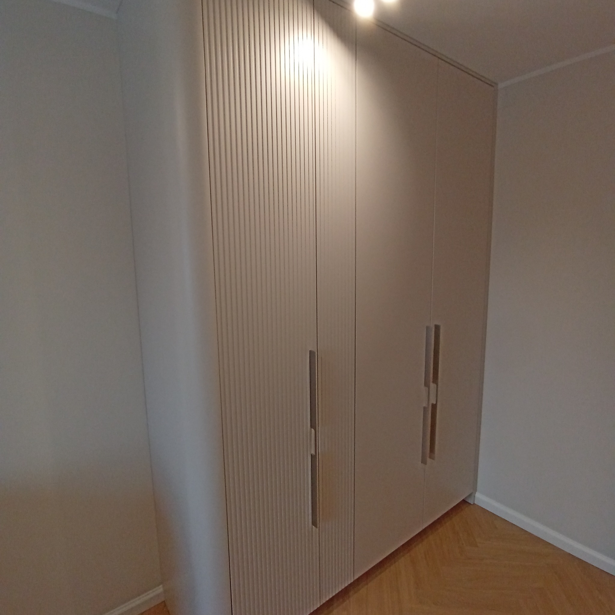 Jasna, wysoka szafa z frezowanymi drzwiami i minimalistycznymi uchwytami, ustawiona na parkiecie w jasnym wnętrzu. Widok z dołu, oświetlenie sufitowe.