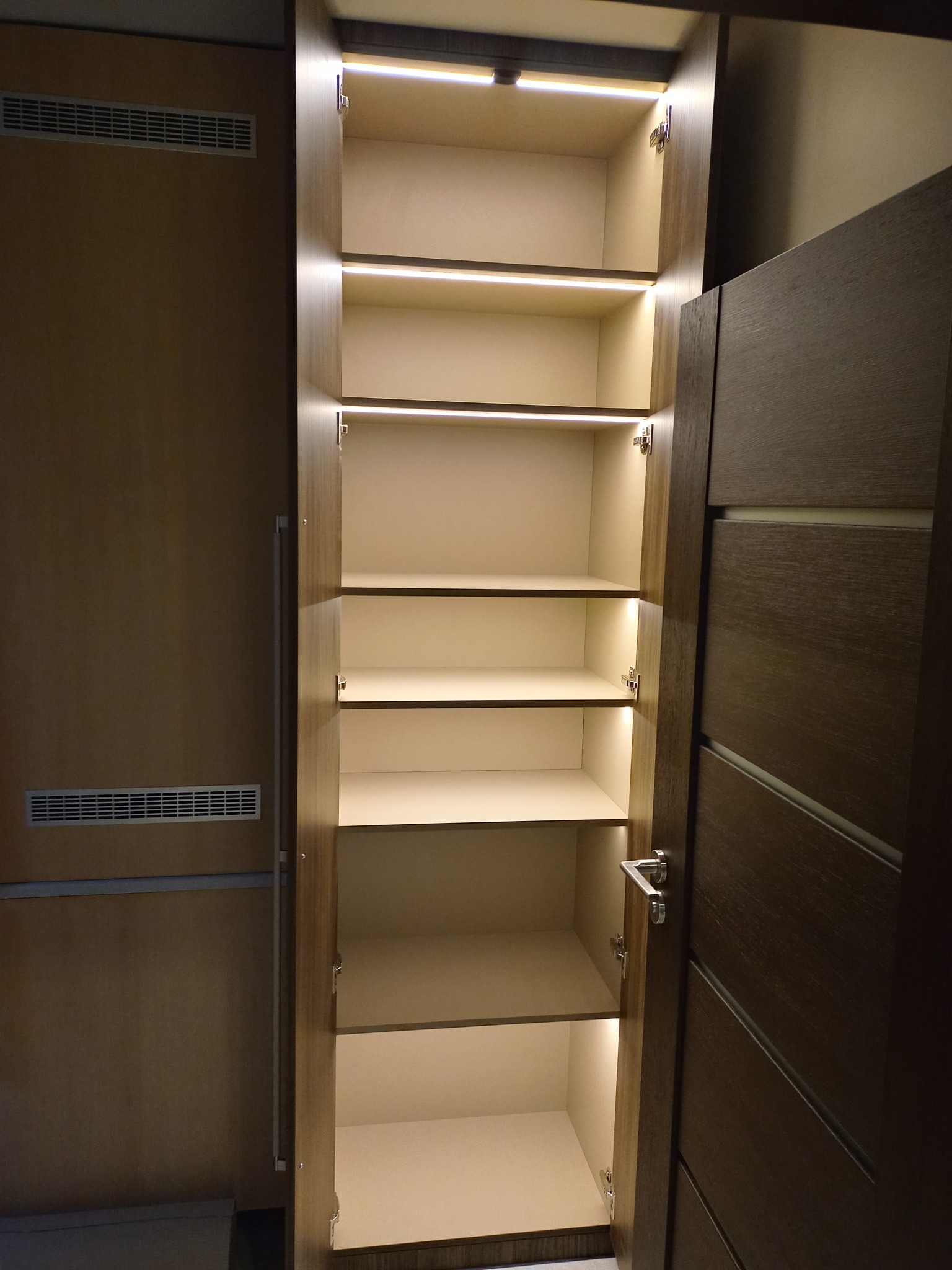 Otwarta, wysoka szafa z półkami i oświetleniem LED, widoczna z bliska. Drzwi szafy w kolorze ciemnego drewna, wnętrze jasne. Obok widoczny fragment ściany z kratką wentylacyjną.