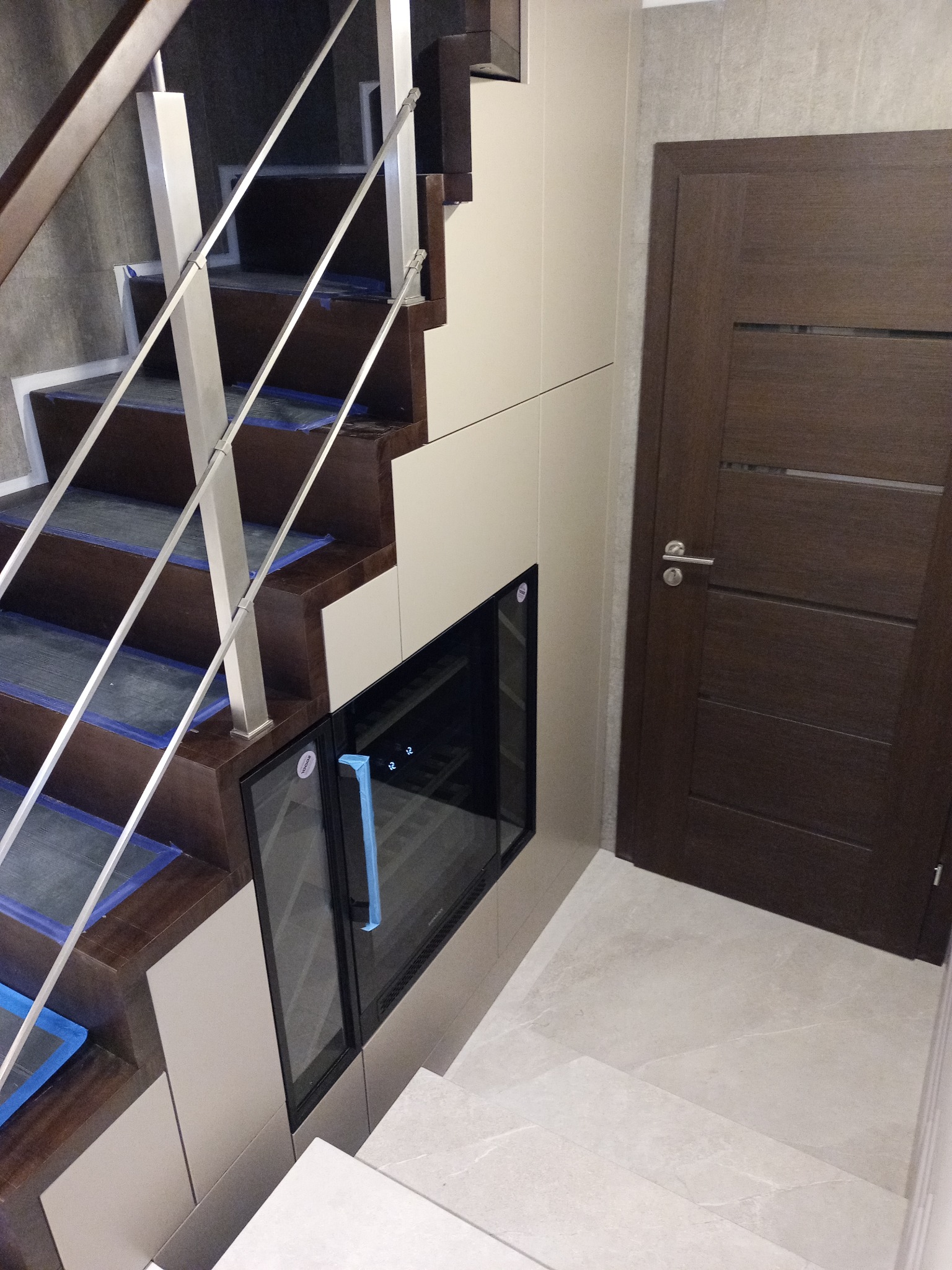 Schody z zabudową meblową i wbudowaną chłodziarką na wino, brązowe stopnie zabezpieczone folią, białe fronty, drzwi wewnętrzne w kolorze wenge.