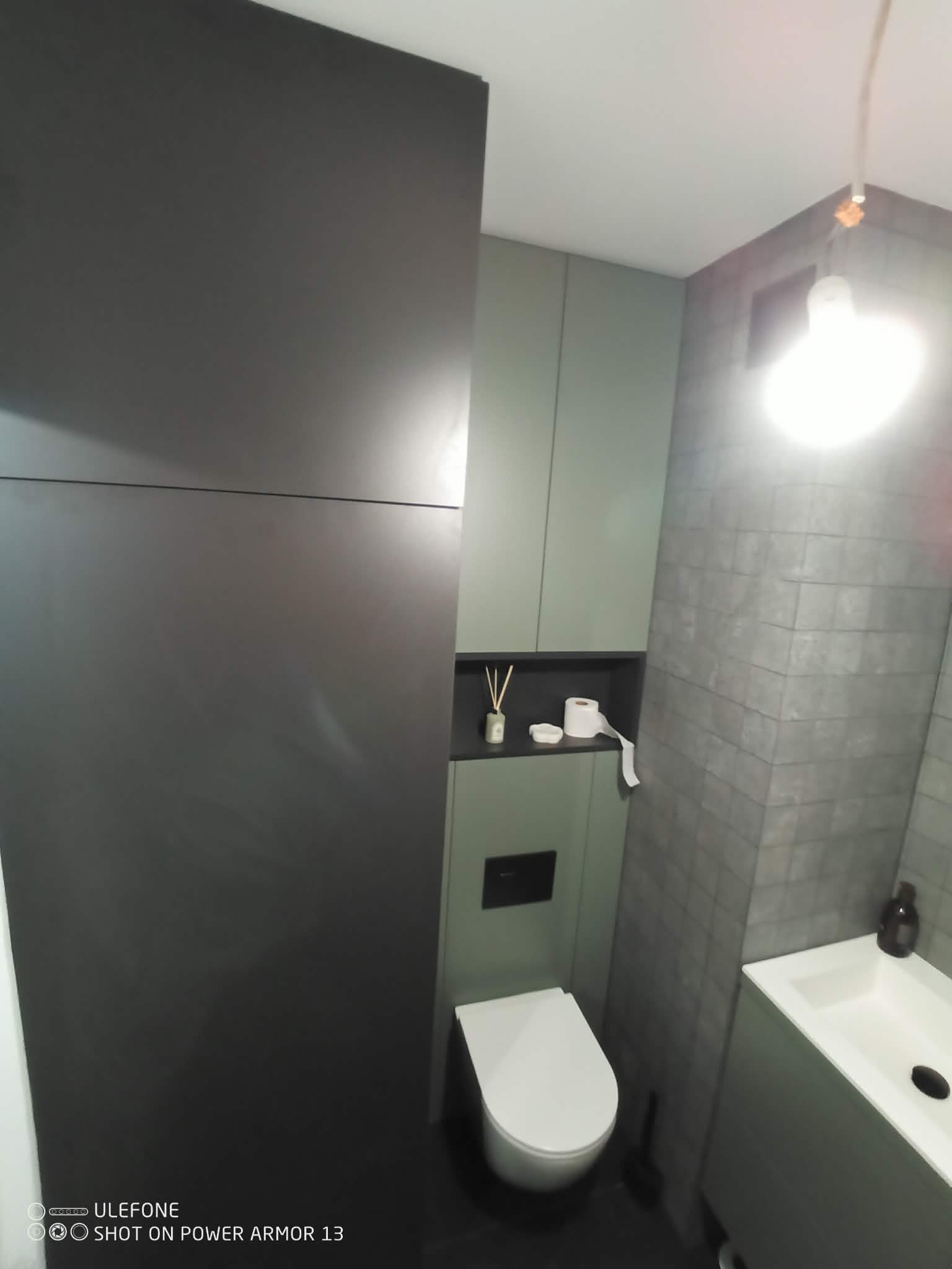 Nowoczesna łazienka z zabudową meblową w odcieniach szarości i zieleni. Wisząca toaleta, umywalka z szafką i wysoka szafa z półkami tworzą spójną całość.